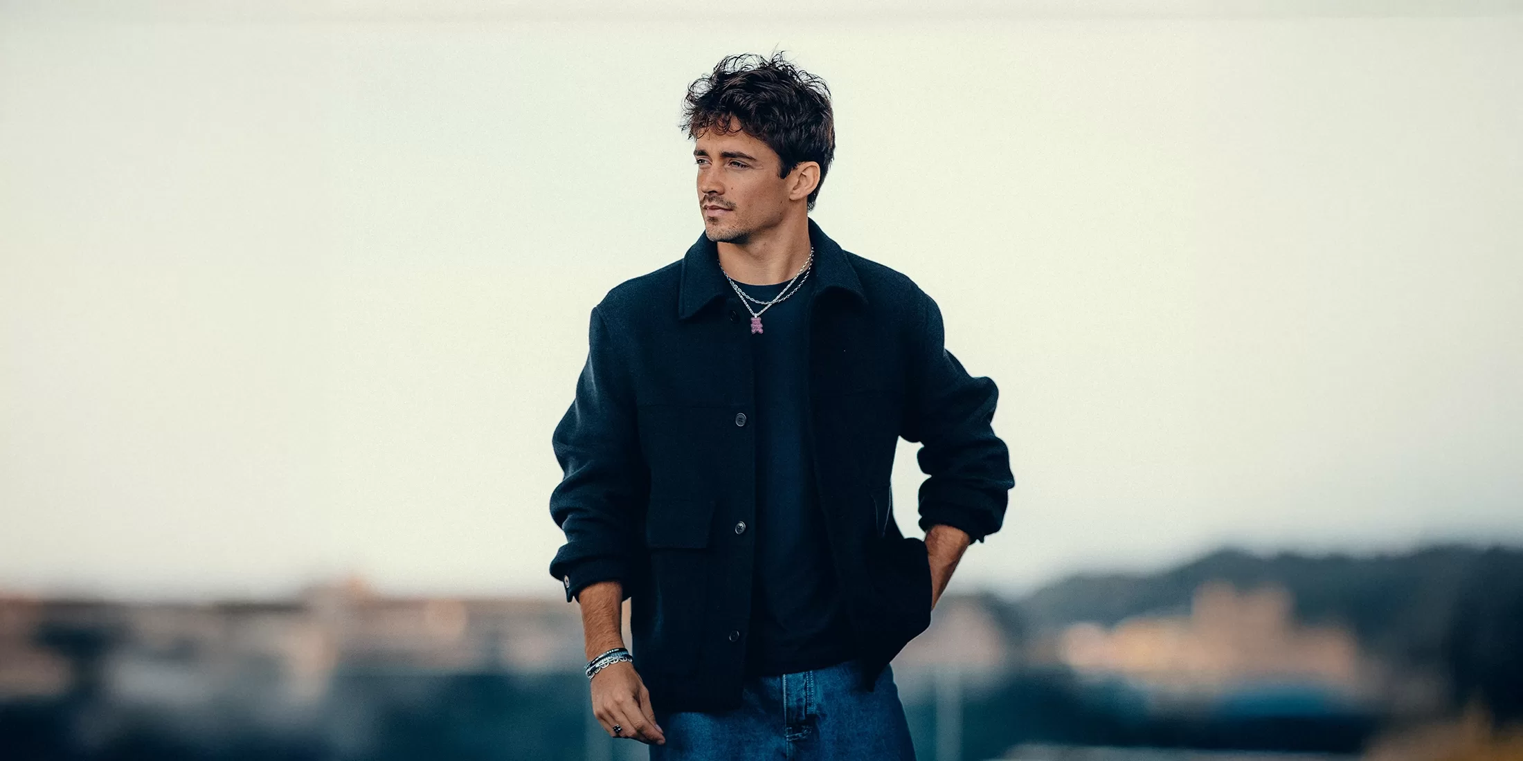 Charles Leclerc - with curly hair stands outdoors wearing a dark jacket, black shirt, and jeans. He has a thoughtful expression, with his right hand in his pocket. The background is blurred, showing a soft, neutral sky and some indistinct structures.