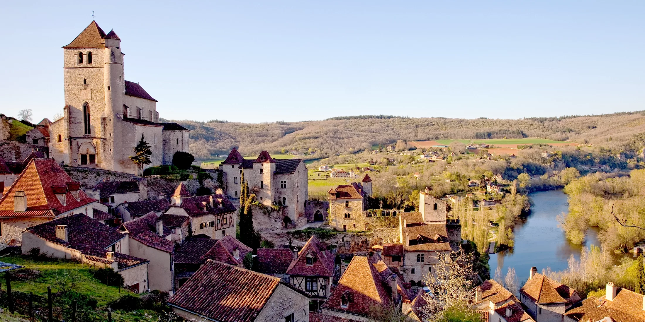 A picturesque medieval village with stone buildings and red-tiled roofs is nestled along a curving river, surrounded by rolling hills and trees under a clear blue sky. A church with a tower overlooks the charming scene.