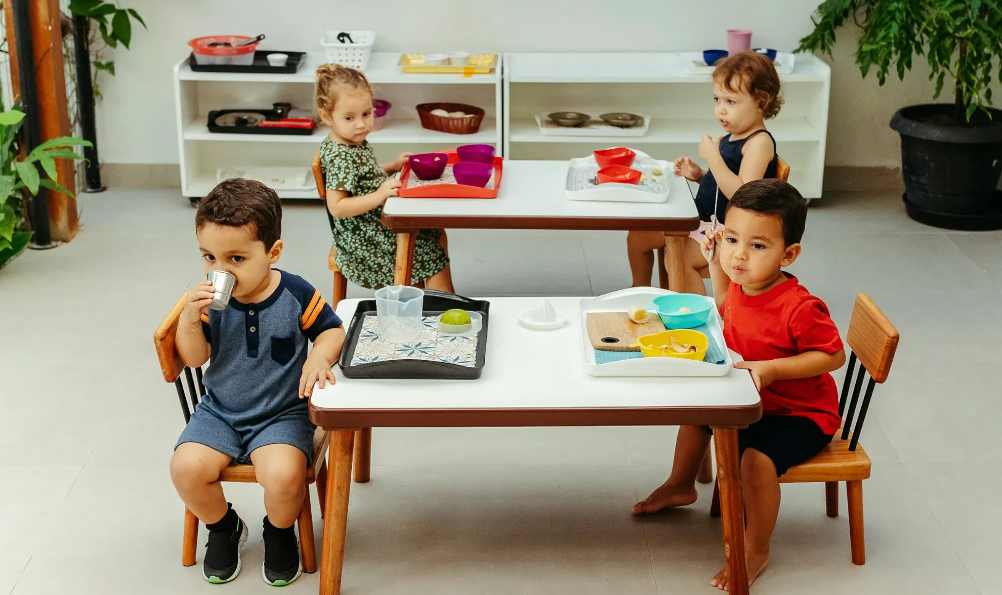 Four young children sit at two small tables with snacks and drinks. They are engaged with their food and drinks. Shelves with various items are in the background, and there are plants around the room.
