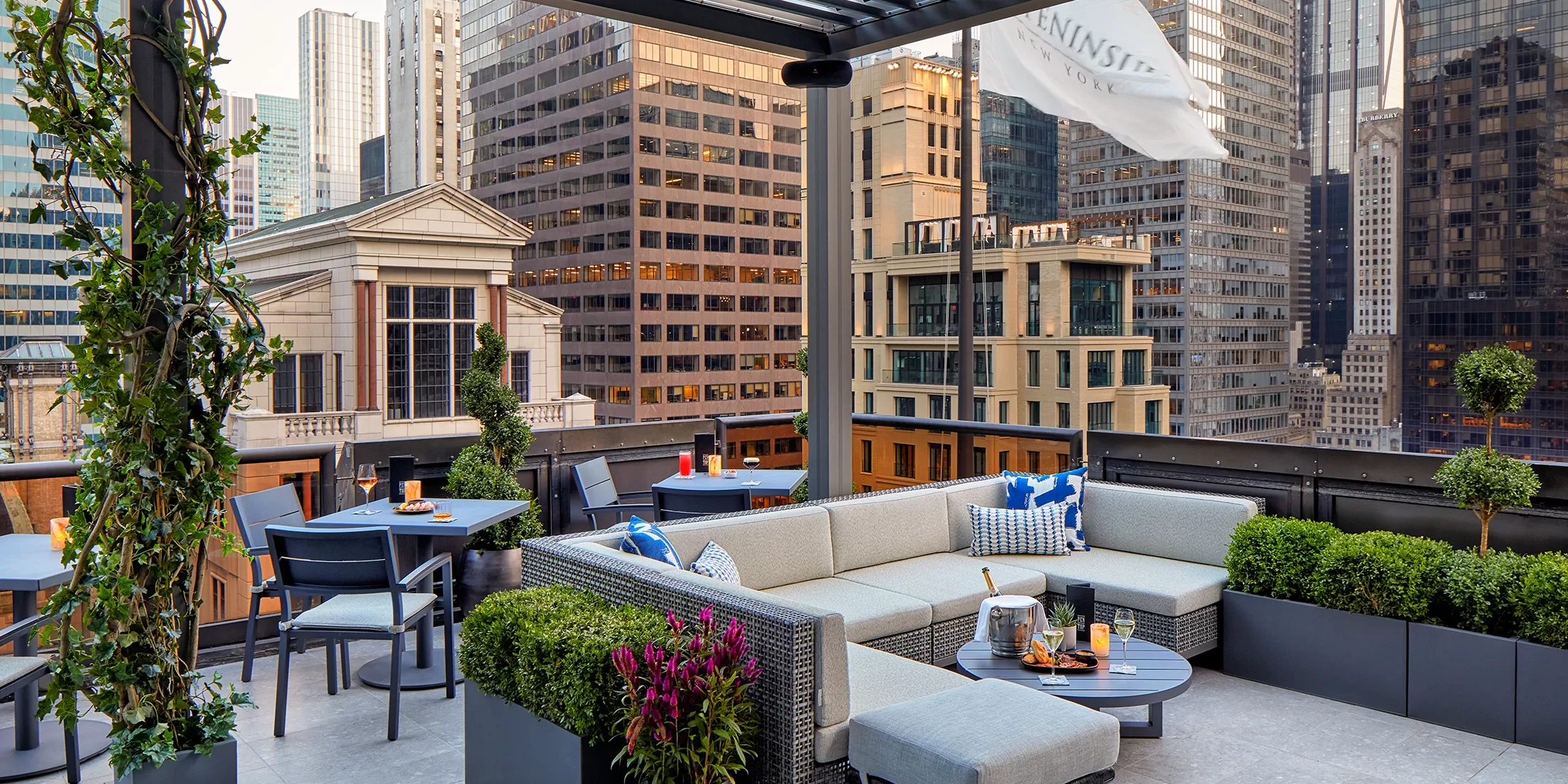 Modern rooftop patio overlooking a cityscape with tall buildings. Features a cozy gray sectional sofa, small tables with chairs, potted plants, and a white flag. Tables have drinks and a champagne bucket. Urban skyline in the background under a clear sky.