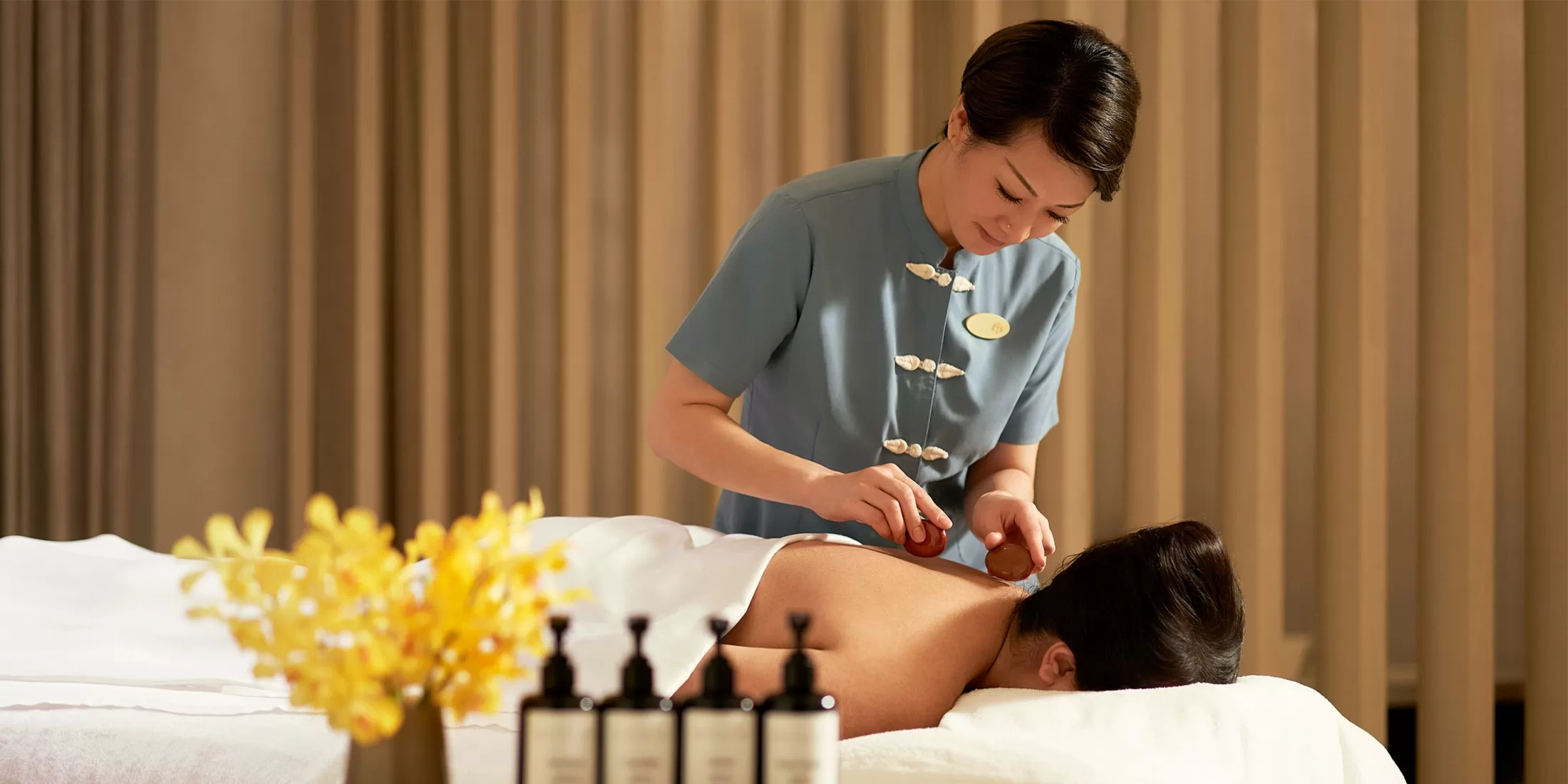 A massage therapist in a blue uniform is performing a treatment on a person lying face down on a massage table. There are bottles of oil in the foreground and yellow flowers nearby. The background features soft, neutral-colored curtains.