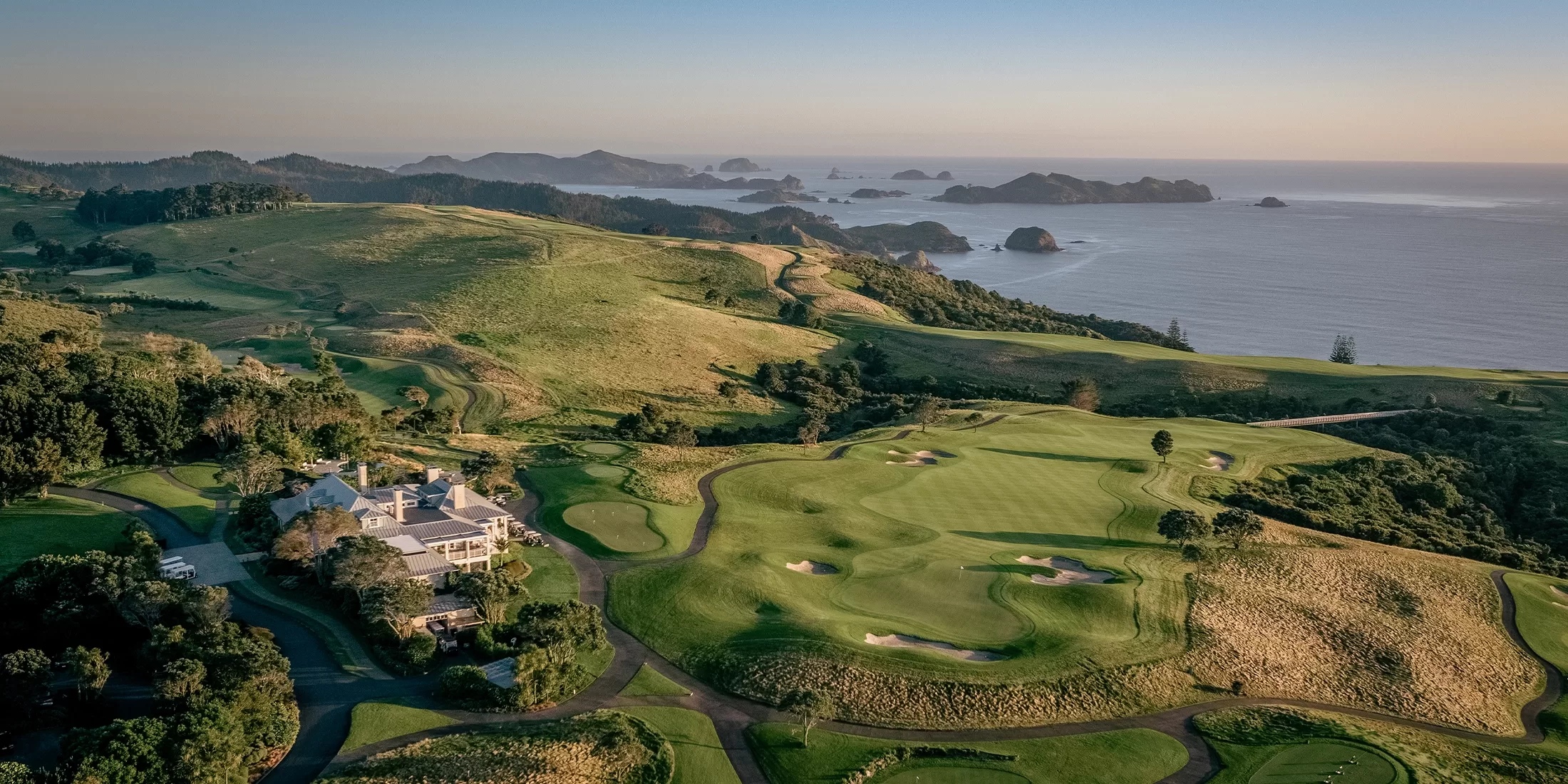 Aerial view of a coastal golf course with lush green fairways and bunkers, surrounded by rolling hills. A large clubhouse is visible on the left. The ocean and small islands are in the background under a clear blue sky.