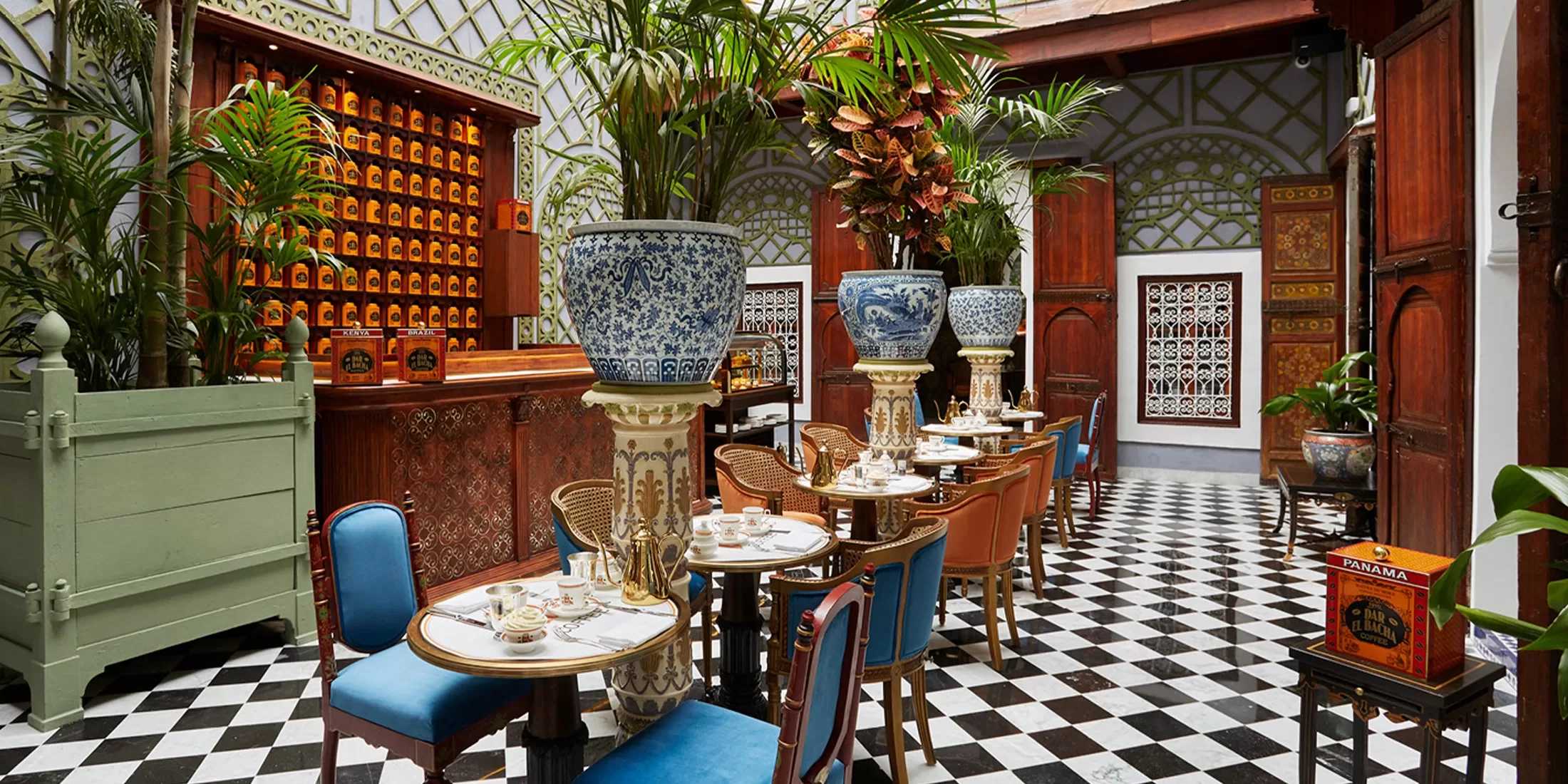 Elegant indoor cafe with black-and-white checkered floor, tall potted plants, blue and gold chairs, small tables set for tea, and ornate wooden decor, including a vintage mail organizer and decorative vases.