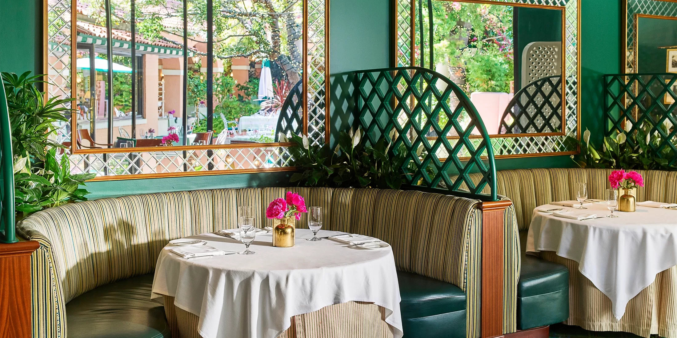 Elegant restaurant interior with green upholstered booths, white tablecloths, gold vases with pink flowers, green lattice dividers, large mirrors, and greenery, with outdoor patio visible through the windows.