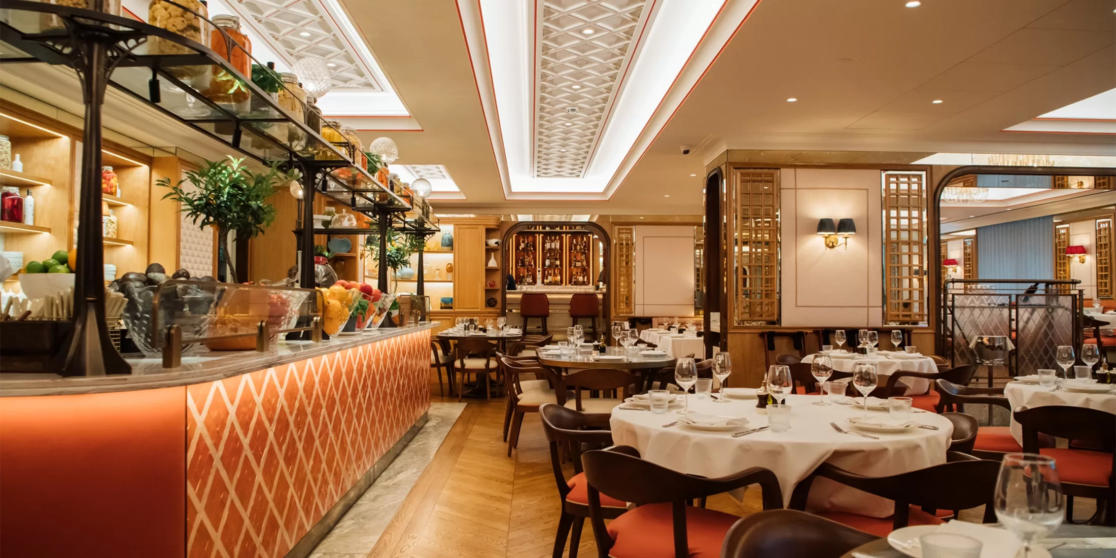 Elegant restaurant interior with round tables set with white cloths and glassware, warm lighting, cushioned chairs, a decorated bar with plants and fruits, and intricate ceiling and wall details.