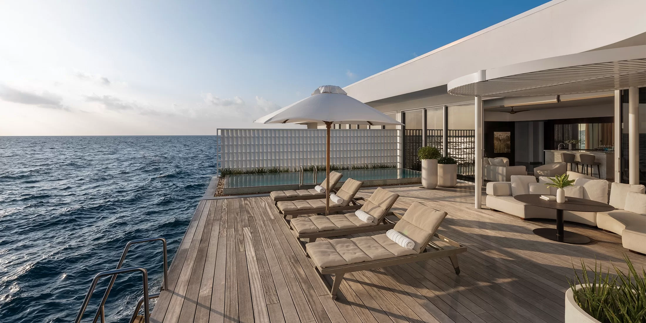 A modern deck with cushioned lounge chairs, a large white umbrella, and an infinity pool overlooks a calm ocean under a clear blue sky, connected to a sleek, open-concept building.