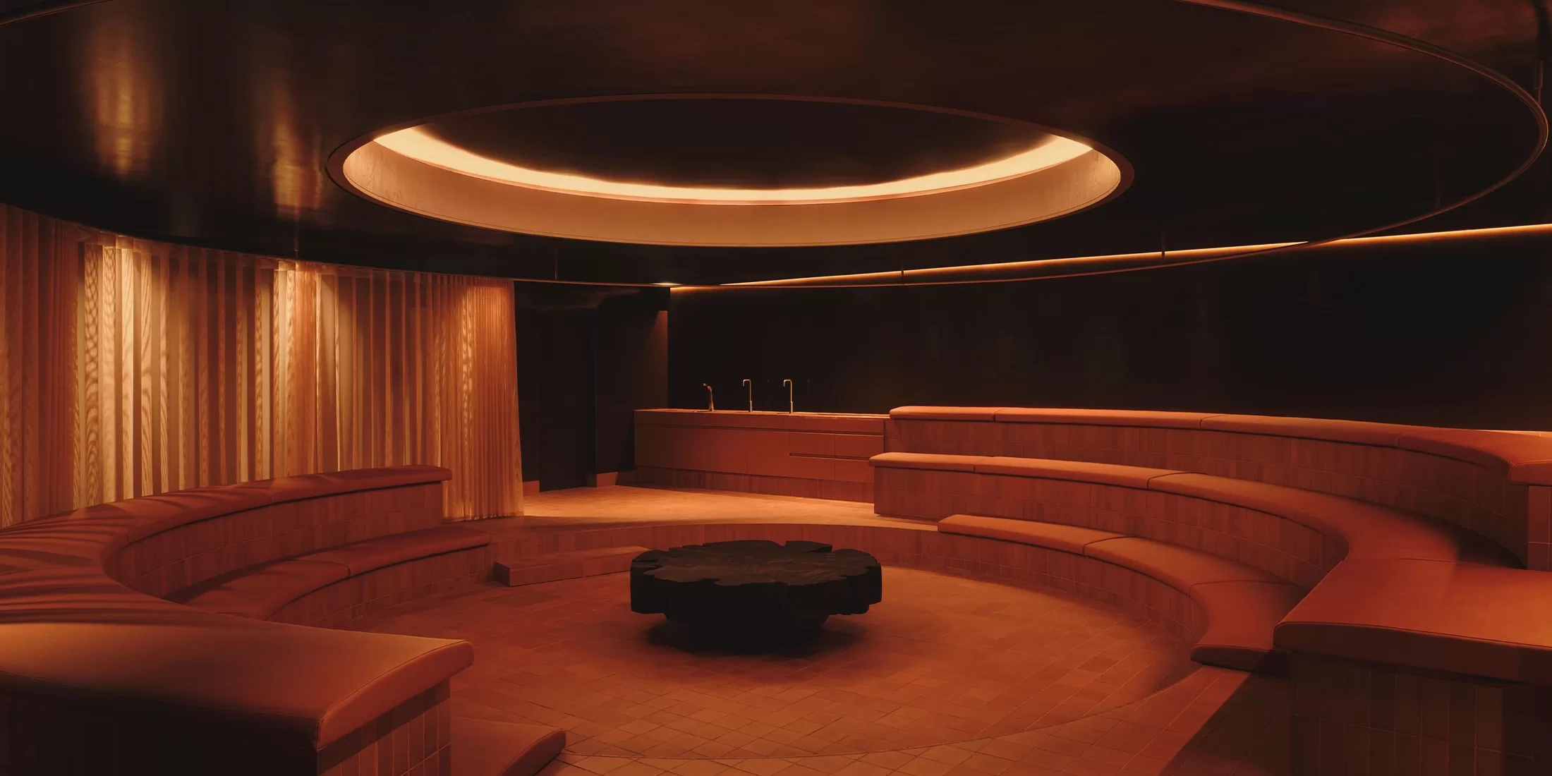 A modern circular sauna room with tiered wooden seating, warm orange lighting, a round ceiling light, and a central black table. The walls and ceiling are dark, creating a cozy, inviting atmosphere.