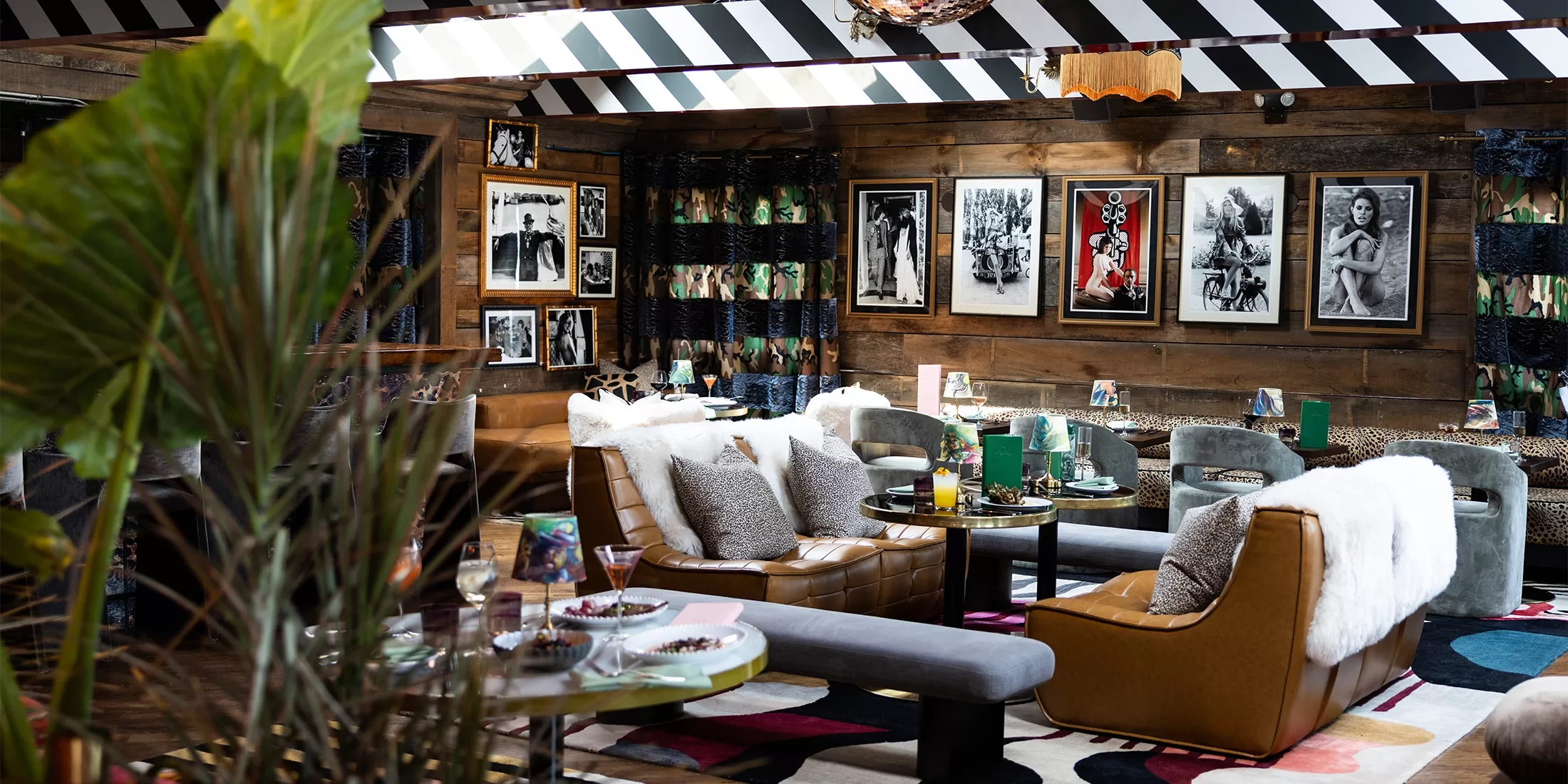 A cozy, stylish lounge with eclectic decor, leather chairs, round tables, colorful rugs, and framed black-and-white photos on wooden walls. Plants and patterned curtains add warmth to the inviting, artistic space.