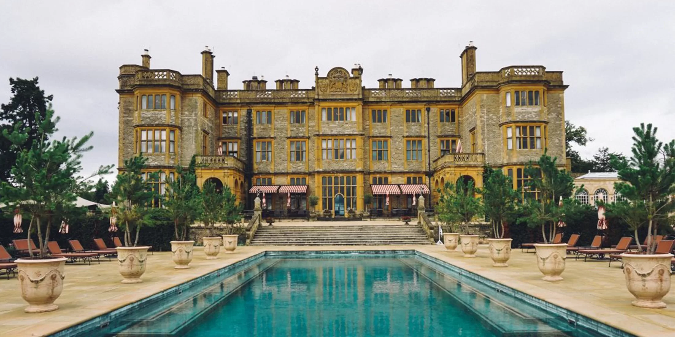 A large, grand manor with ornate architecture stands behind a rectangular outdoor pool, surrounded by potted plants, loungers, and trees under a cloudy sky.