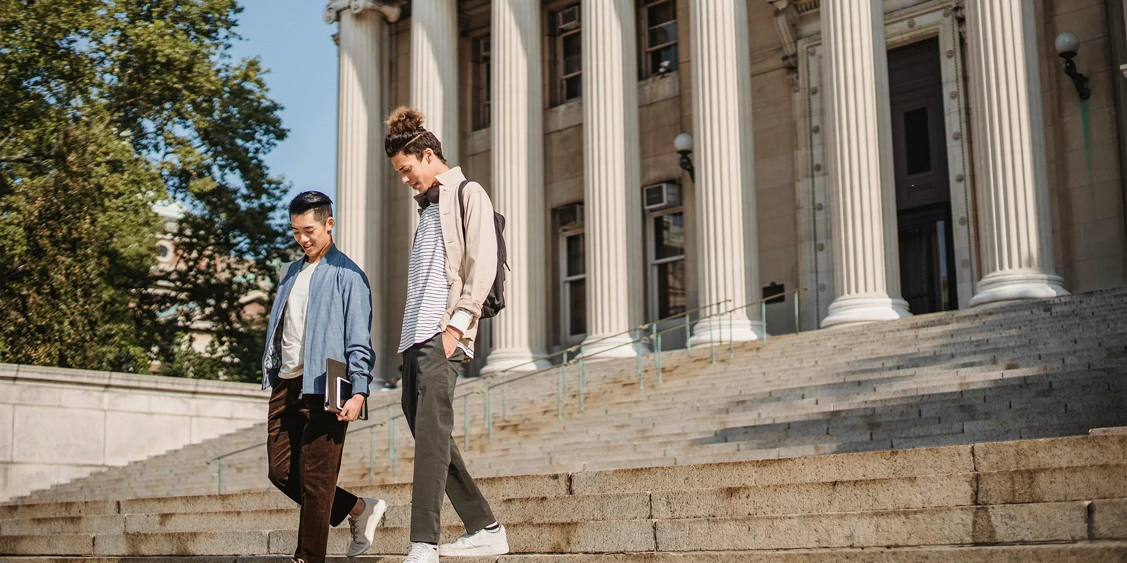 Two students walk down stone steps outside a large building with tall columns, holding books and backpacks, smiling and talking on a sunny day.