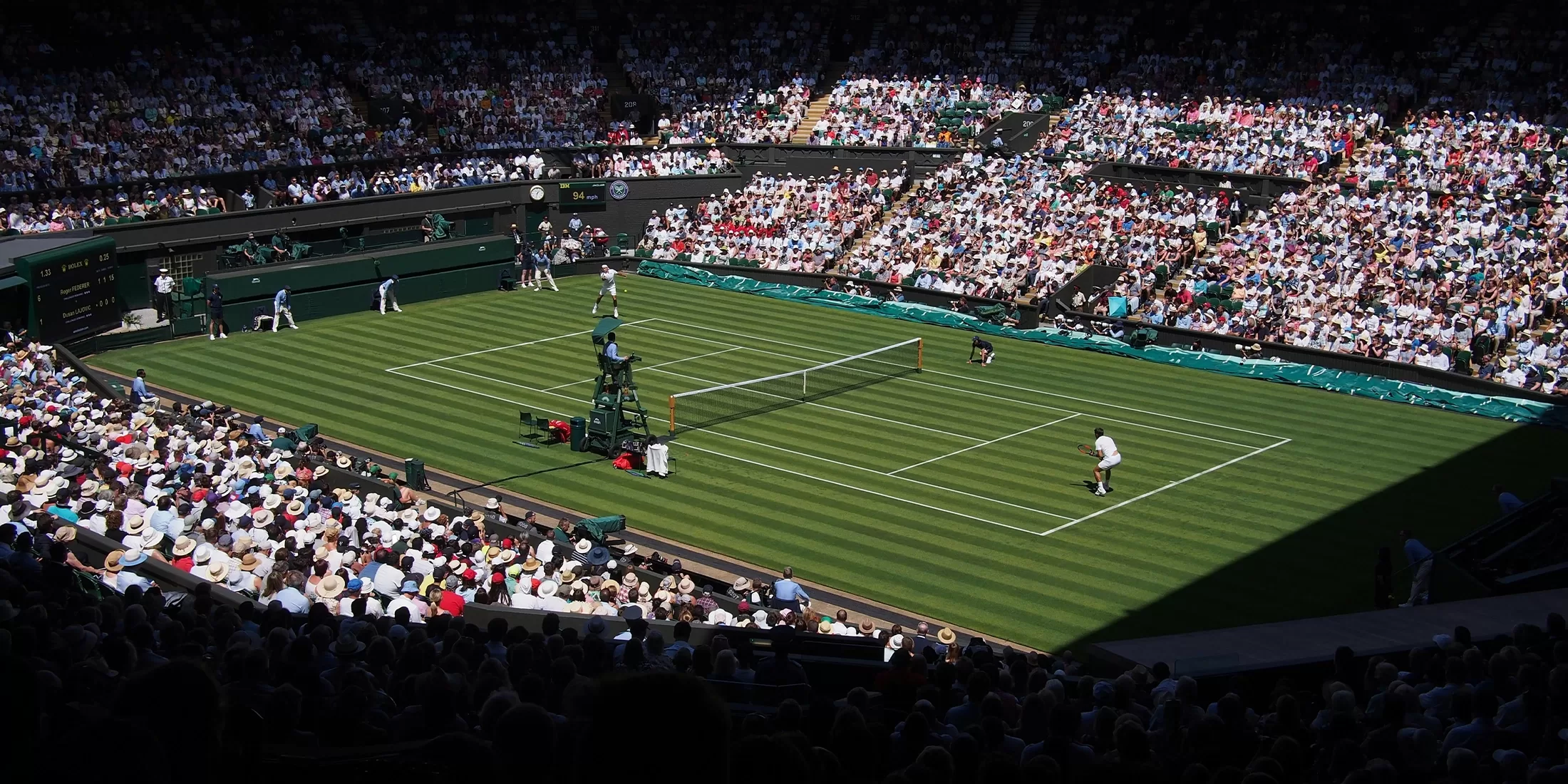 A large crowd watches a tennis match on a grass court in a stadium. Two players are on the court, officials and ball kids are present, and sunlight partially illuminates the audience and playing area.