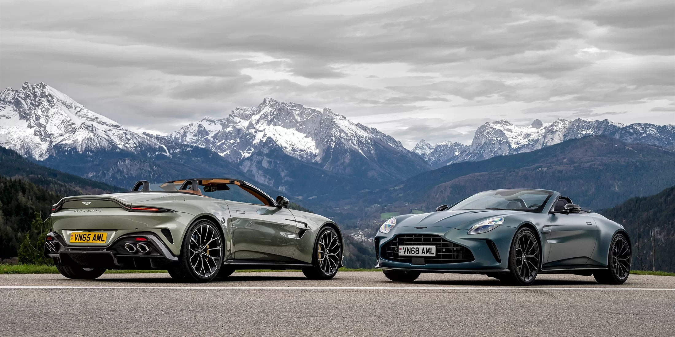 Two sleek, sporty convertibles are parked on a mountain road with snow-capped peaks and dramatic clouds in the background. The cars face in opposite directions, highlighting their elegant designs and glossy finishes.