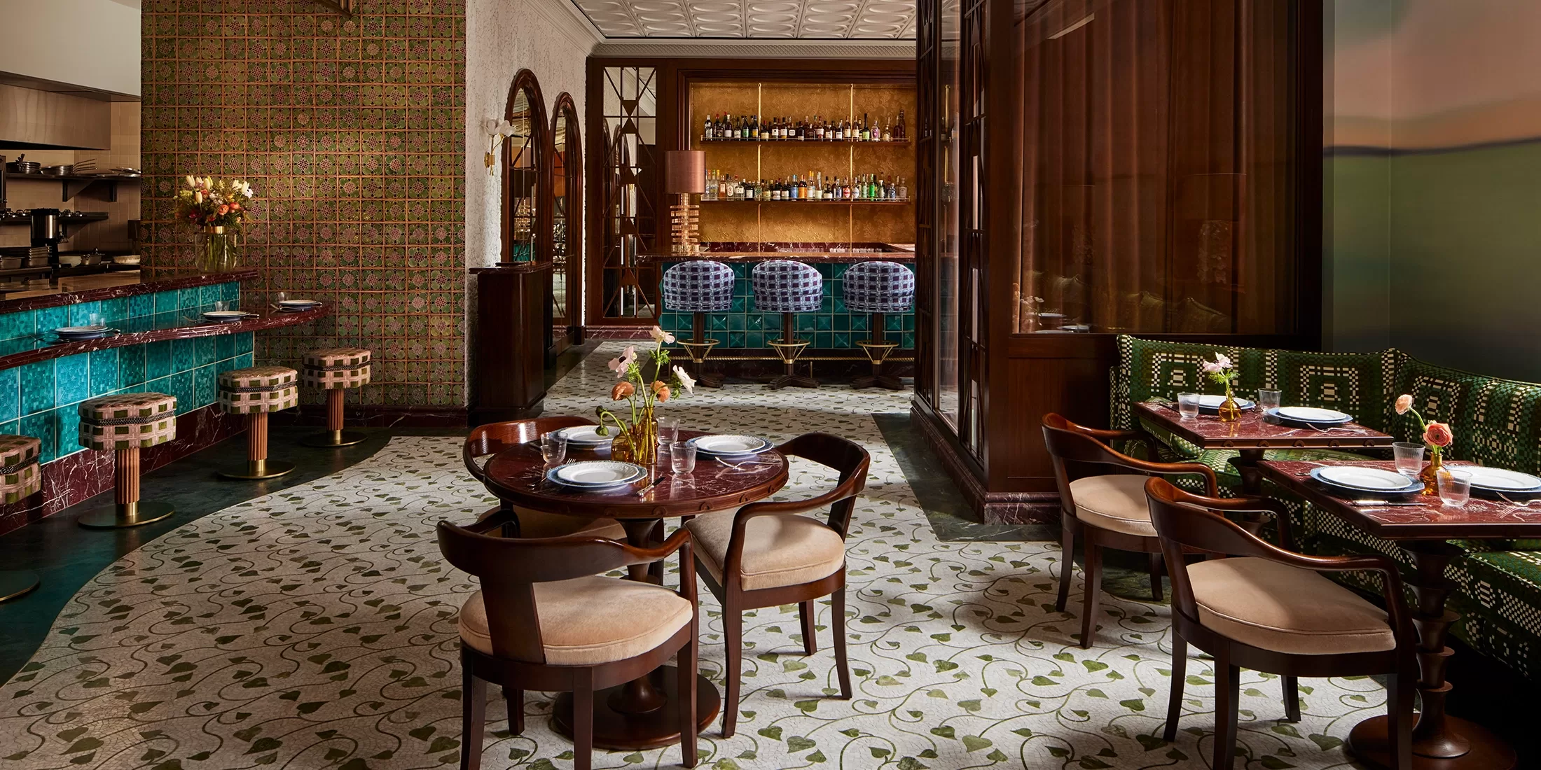 Elegant restaurant interior with wooden tables and chairs, green patterned booths, a turquoise tiled bar, and decorative lighting; the space features a warm, inviting atmosphere with luxurious and artistic design elements.