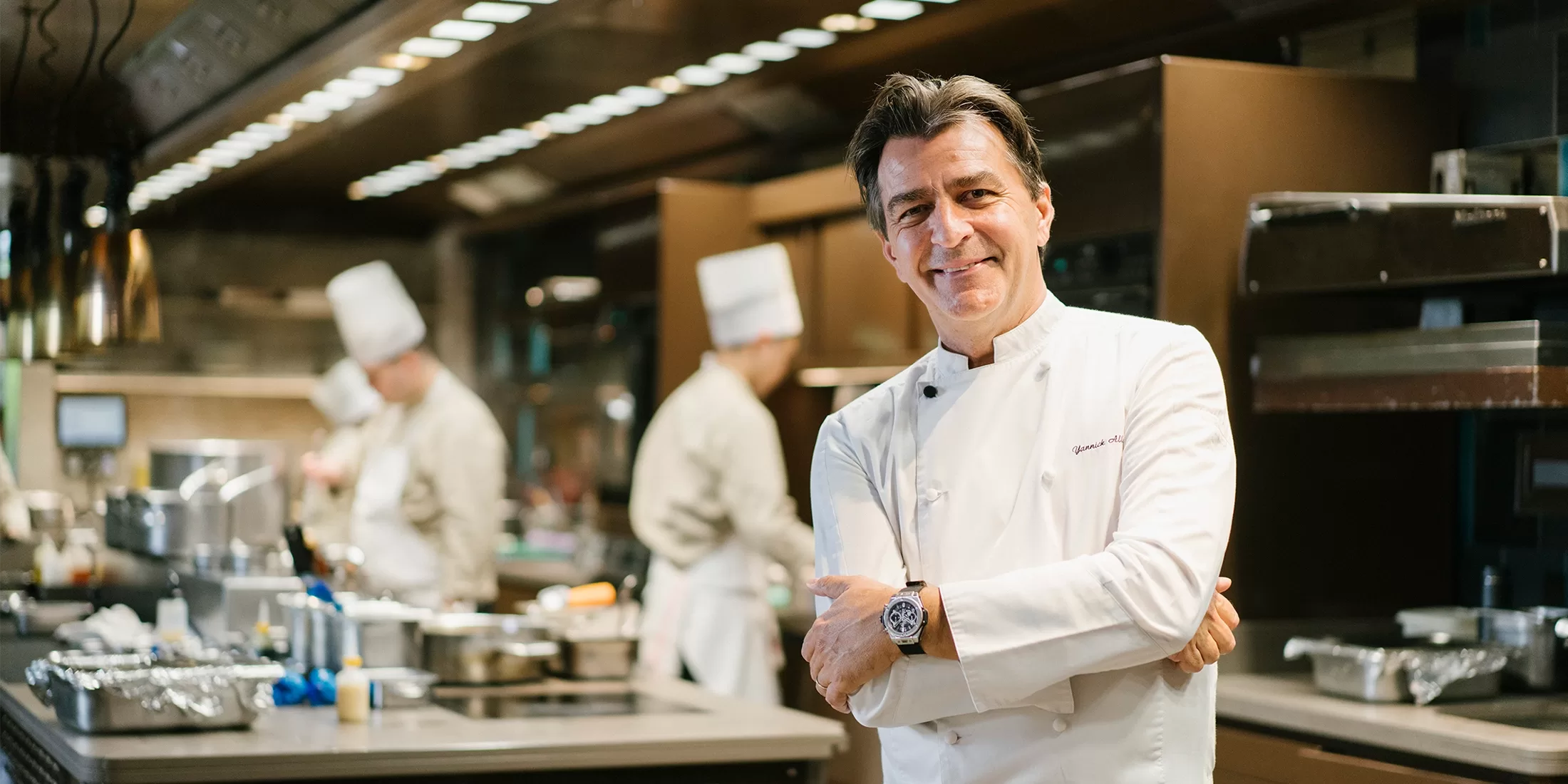 A smiling chef in a white uniform stands with arms crossed in a professional kitchen, while two other chefs work in the background.
