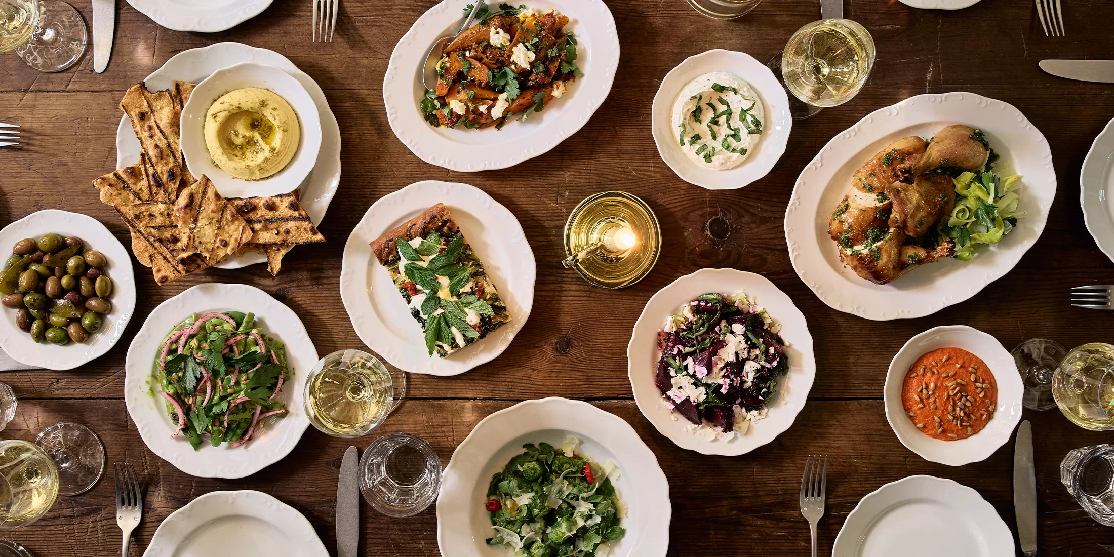 A wooden table set for a meal with white plates and bowls holding salads, roasted vegetables, dips, bread, olives, and glasses of white wine. Cutlery and glasses are arranged around the dishes.
