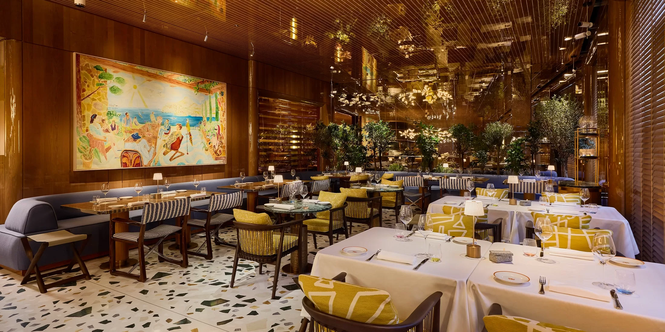 Upscale restaurant interior with elegant tables set for dining, yellow and white chairs, a large colorful mural on the wooden wall, potted trees, and warm ambient lighting reflected on shiny ceiling panels.