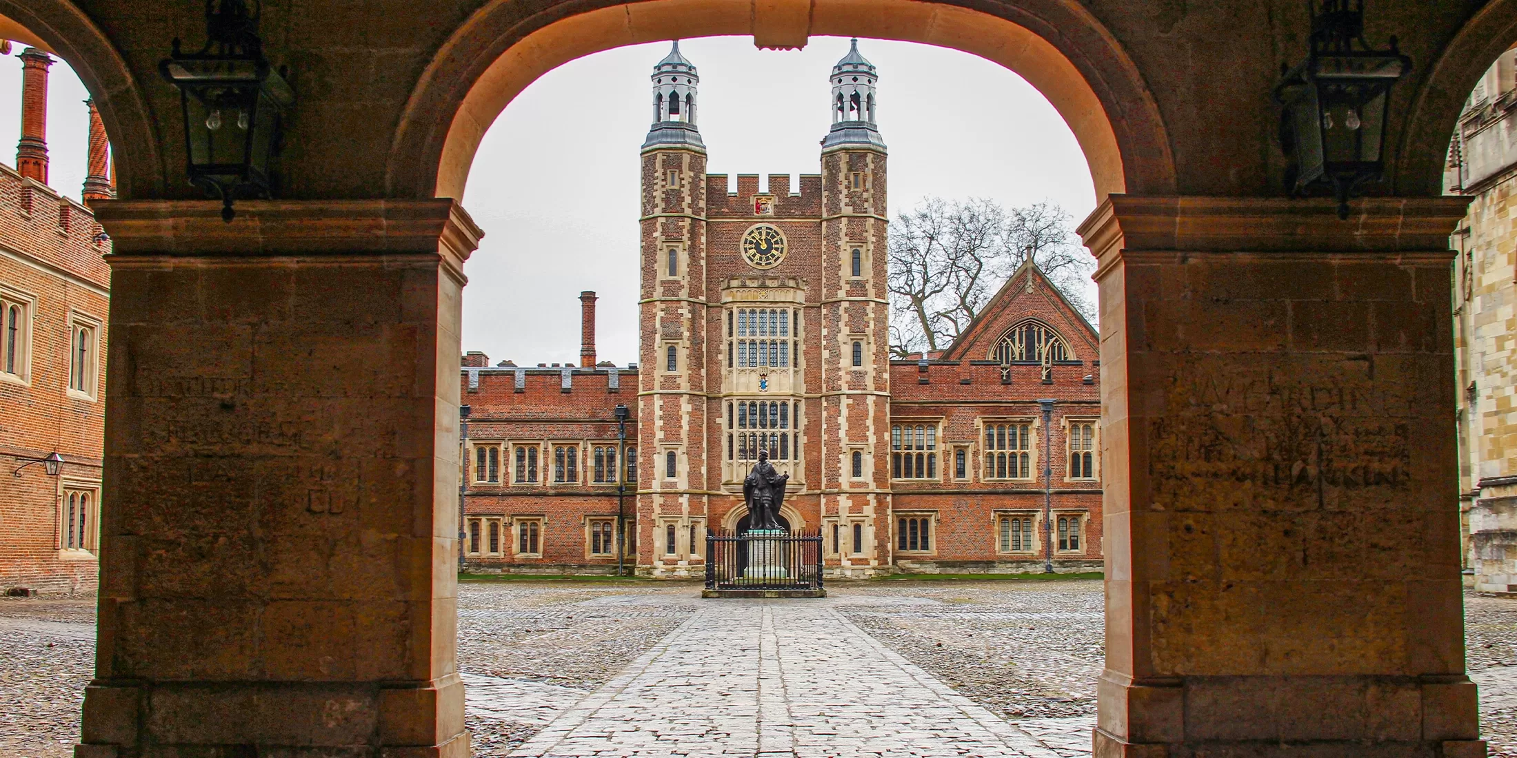 View through a stone archway of a historic brick building with a clock tower, tall chimneys, and a statue in the courtyard, under a cloudy sky.