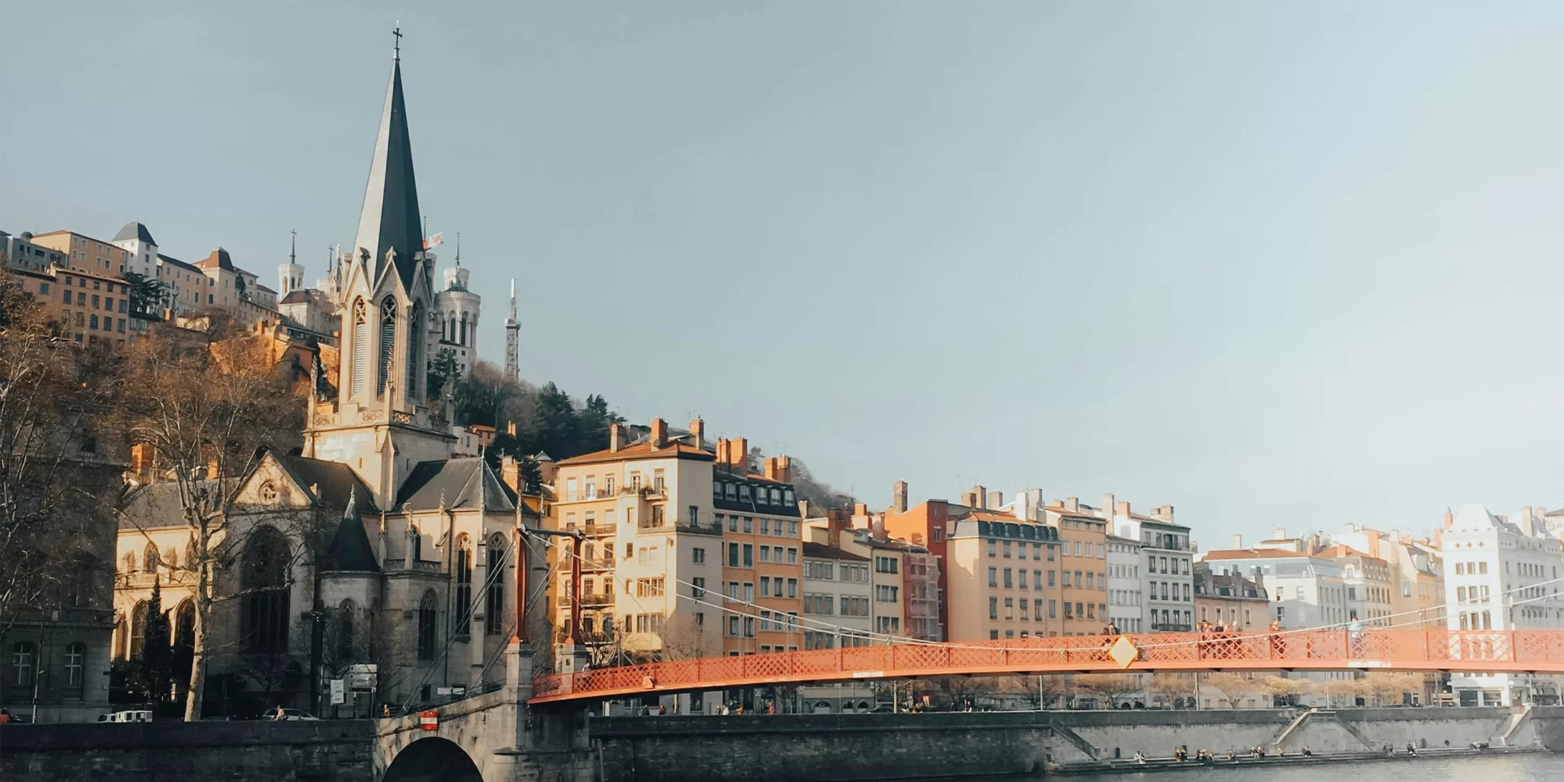 A view of a riverside cityscape with colorful old buildings, a church with a tall steeple, and a red pedestrian bridge crossing the river under a clear sky.