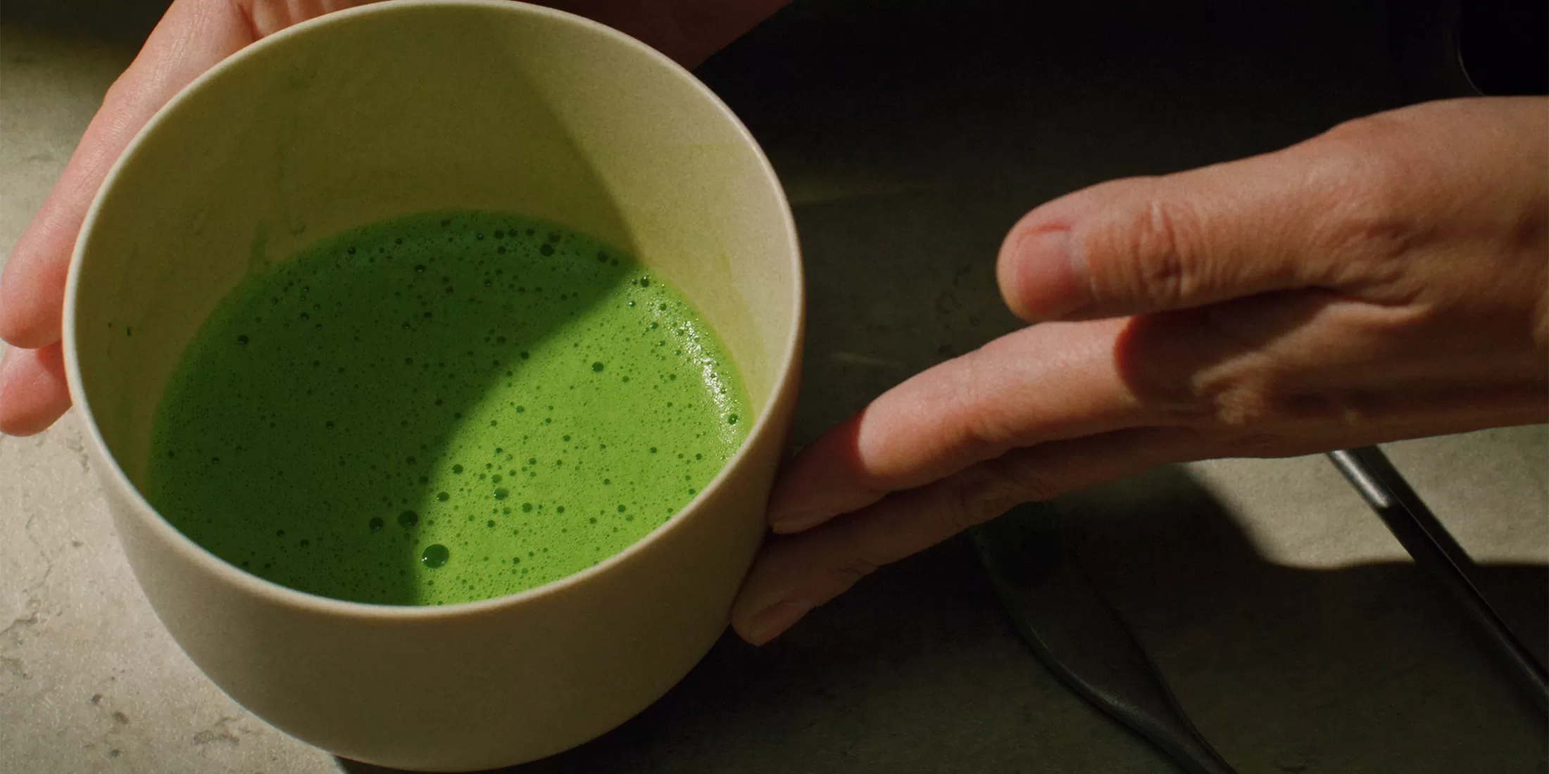 A hand holds a light-colored cup filled with a bright green, frothy liquid on a dark surface.