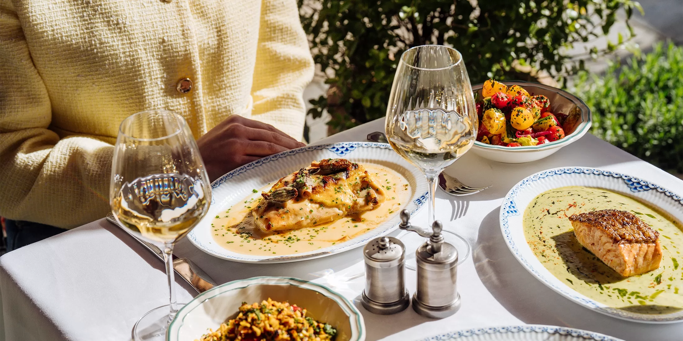 A table set outdoors with plates of gourmet food, including fish fillets in sauce, roasted potatoes with vegetables, and wine glasses. A person in a yellow jacket sits at the table amid greenery.