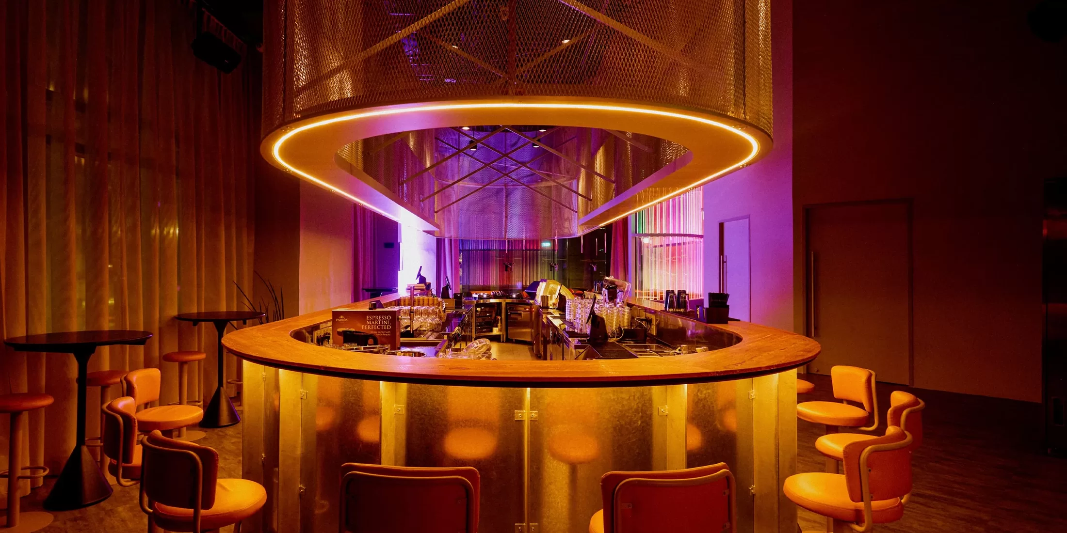 A modern bar with warm orange and purple lighting, round wooden counter, yellow chairs, and a large circular overhead light fixture, creating a stylish and inviting atmosphere.