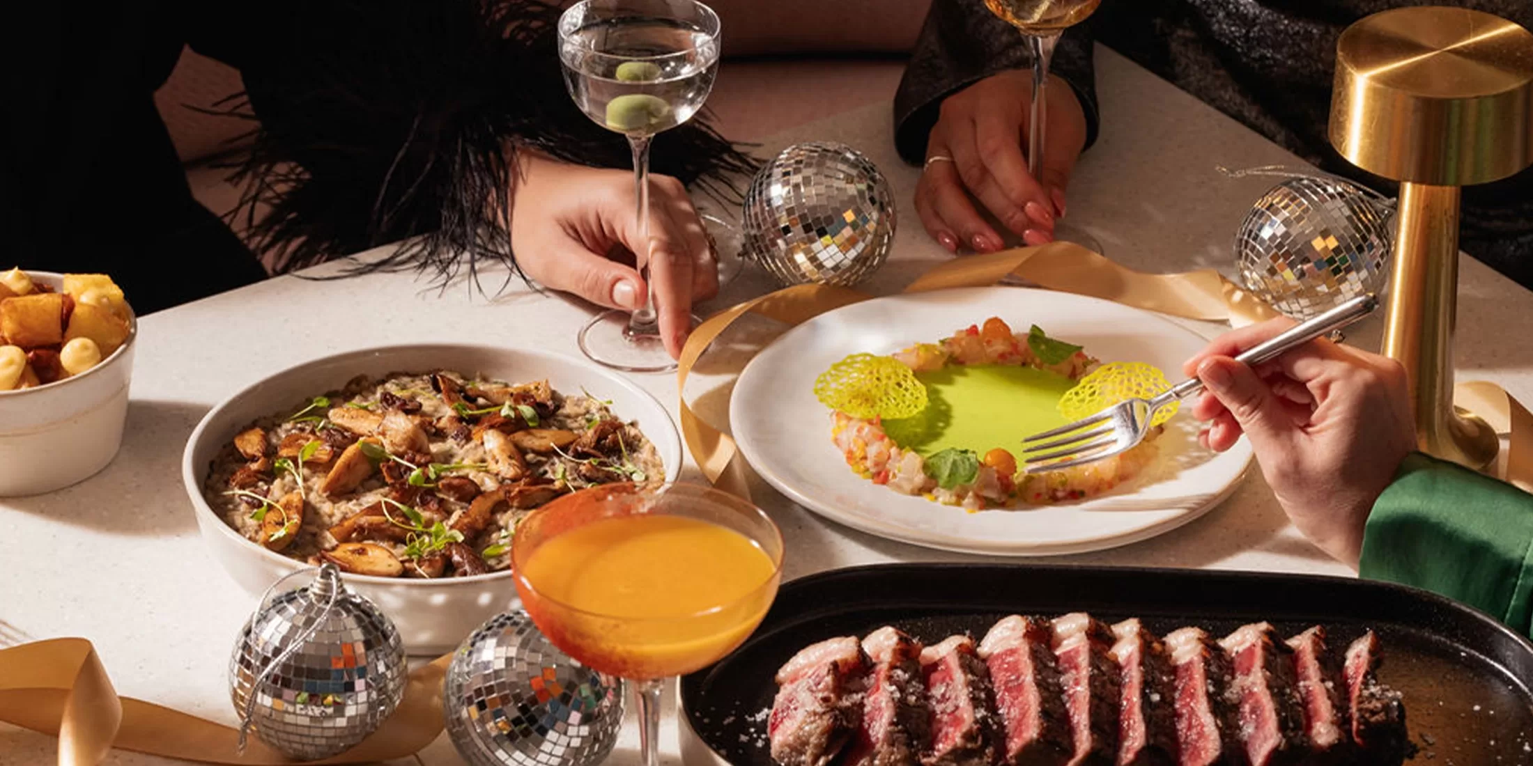 A festive table setting with gourmet dishes, cocktails, golden ribbon, and small disco balls. Three people’s hands are visible, reaching for food and drinks, creating a celebratory atmosphere.