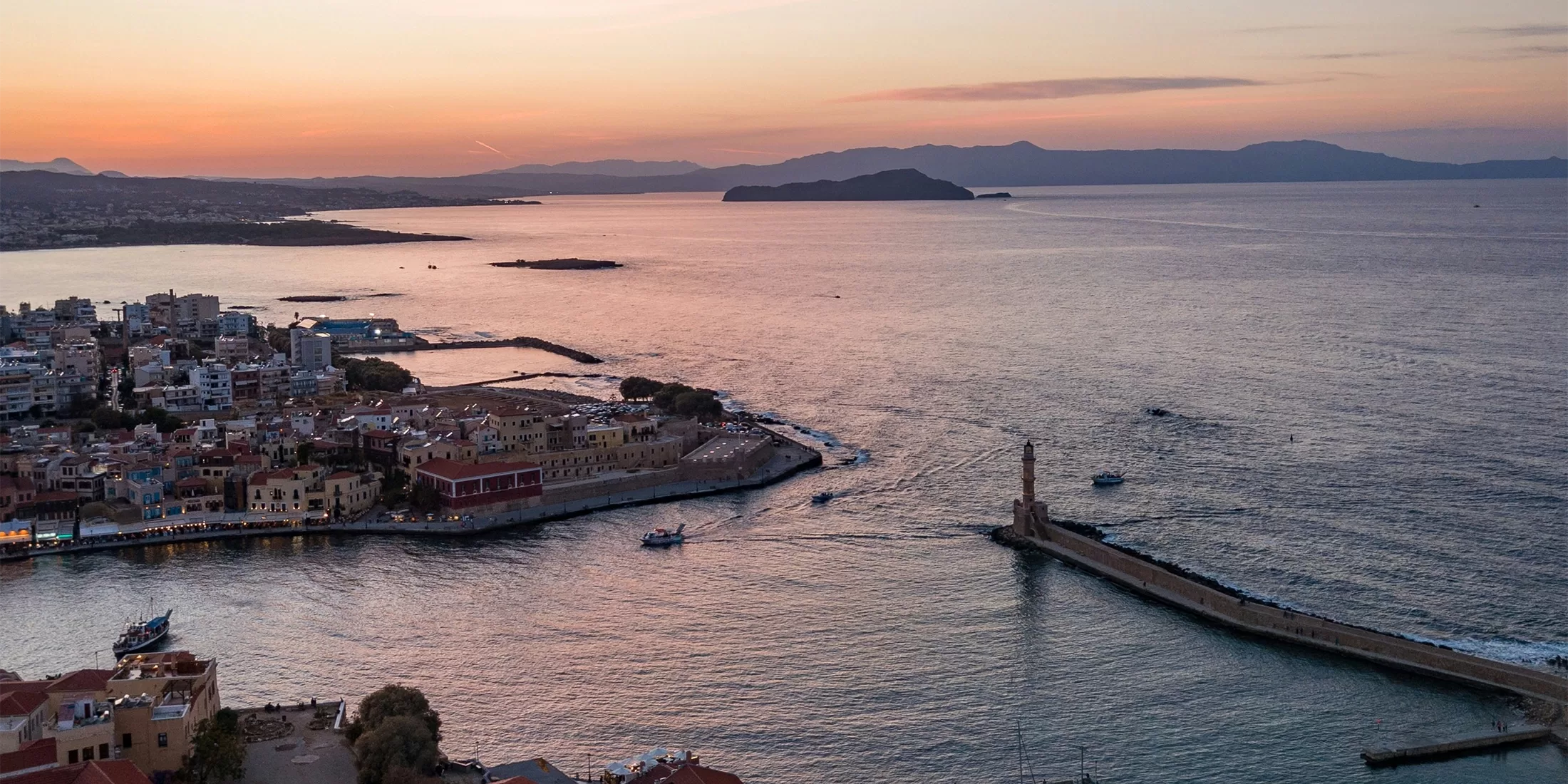 A coastal town at sunset with calm waters, buildings along the shore, a prominent lighthouse on a long pier, and mountains in the background under a colorful sky.