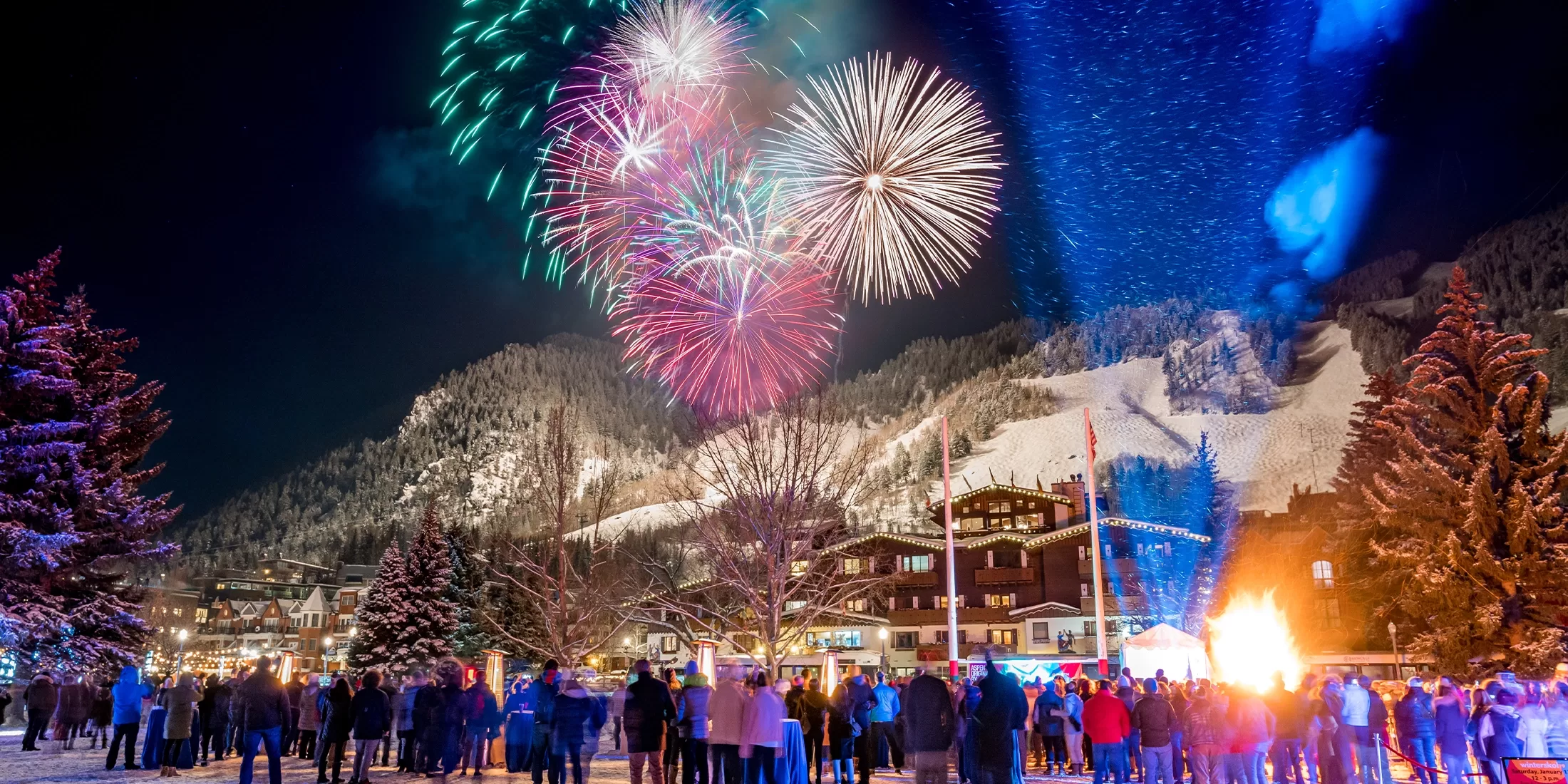 A crowd gathers outdoors on a snowy winter night, watching colorful fireworks burst in the sky above a mountain town with snow-covered trees and buildings.