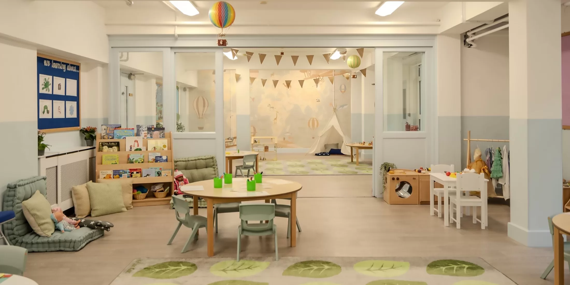 A bright, tidy preschool classroom with round tables, small chairs, bookshelves, toys, and a play tent in the back. Colorful decorations and soft furnishings create a welcoming, child-friendly atmosphere.