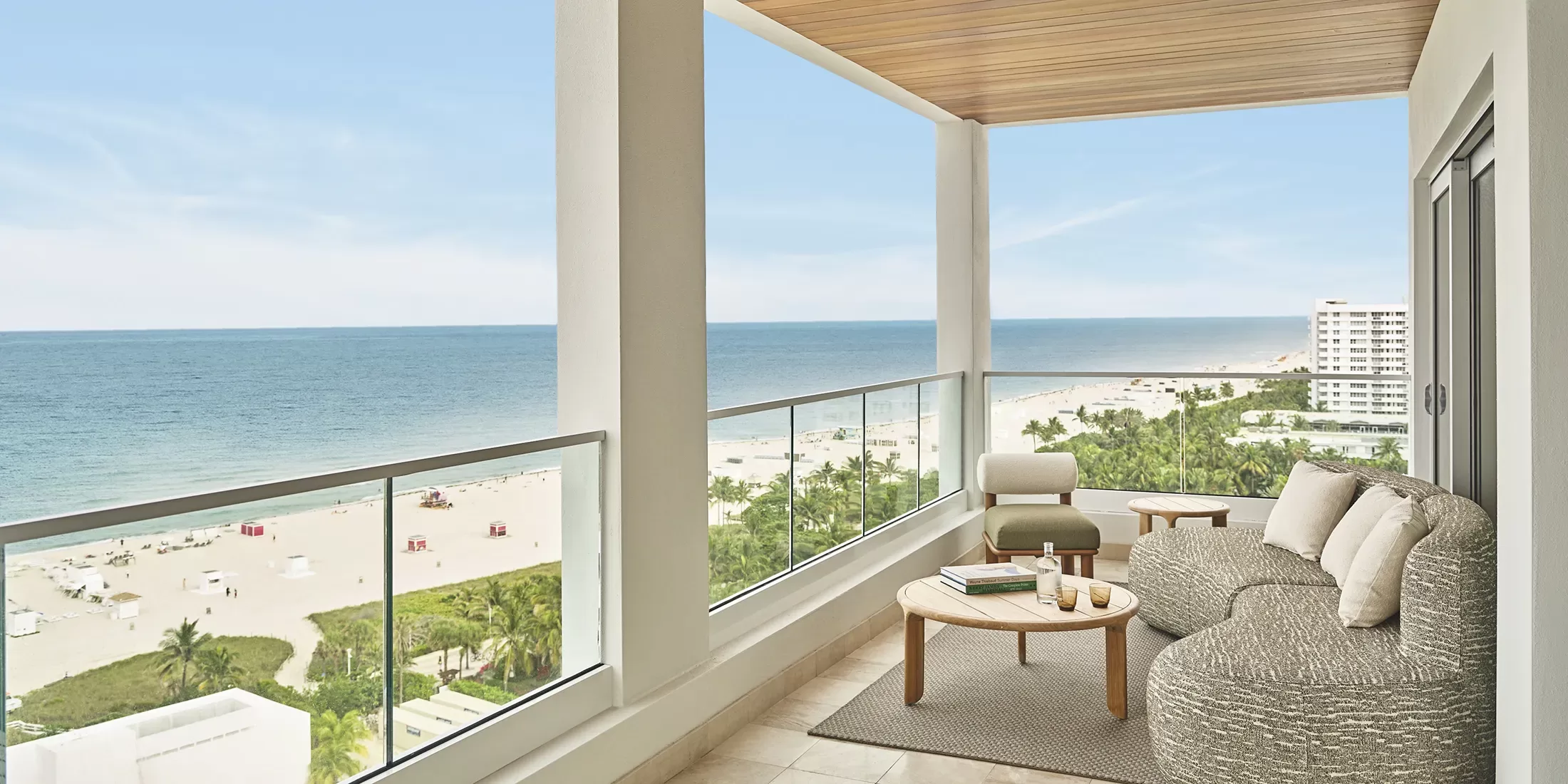 A modern balcony with cozy seating and a table overlooks a sandy beach and the ocean, with clear blue skies and a few distant buildings visible along the shoreline.