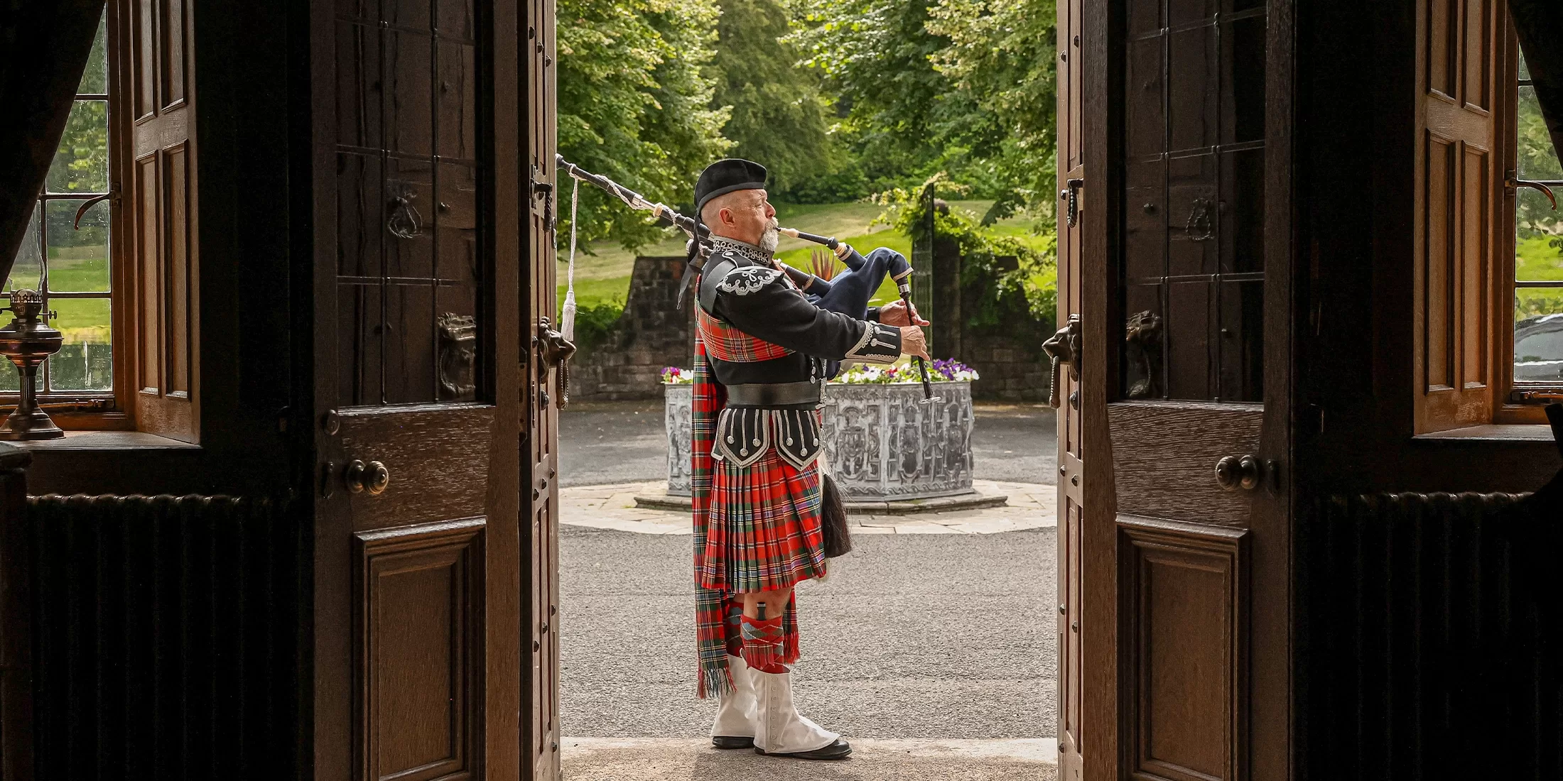 A man in traditional Scottish attire plays bagpipes outside, framed by open wooden doors, with greenery and a stone structure visible in the background.