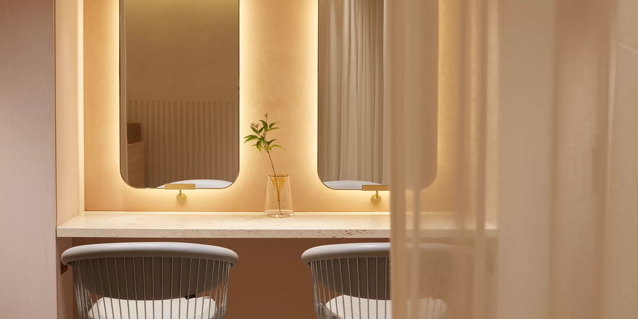 Two sleek chairs face a marble vanity with two large, softly lit mirrors. A single glass vase with a green branch sits between the mirrors, creating a minimalistic and calm atmosphere.