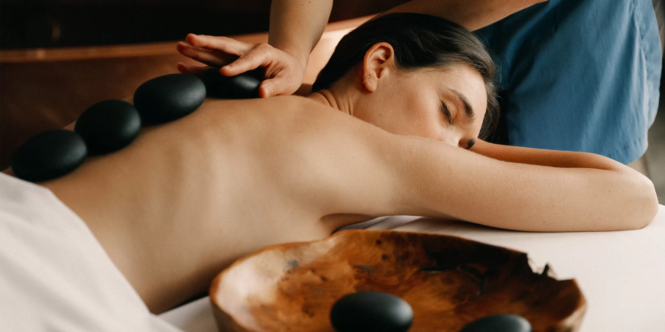 A woman lies face down on a massage table with eyes closed, receiving a hot stone massage. Several smooth black stones are placed along her bare back, and a wooden bowl with more stones is nearby.