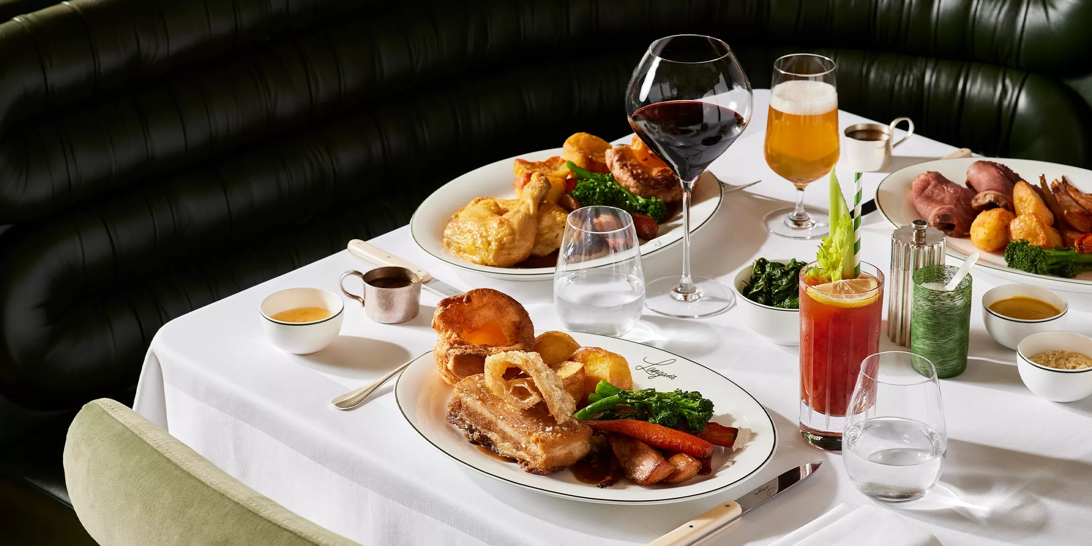 A table set for two with plates of roast dinners, Yorkshire puddings, vegetables, and gravy. Drinks include red wine, beer, a cocktail, and water. The table is covered with a white cloth and set in a green leather booth.