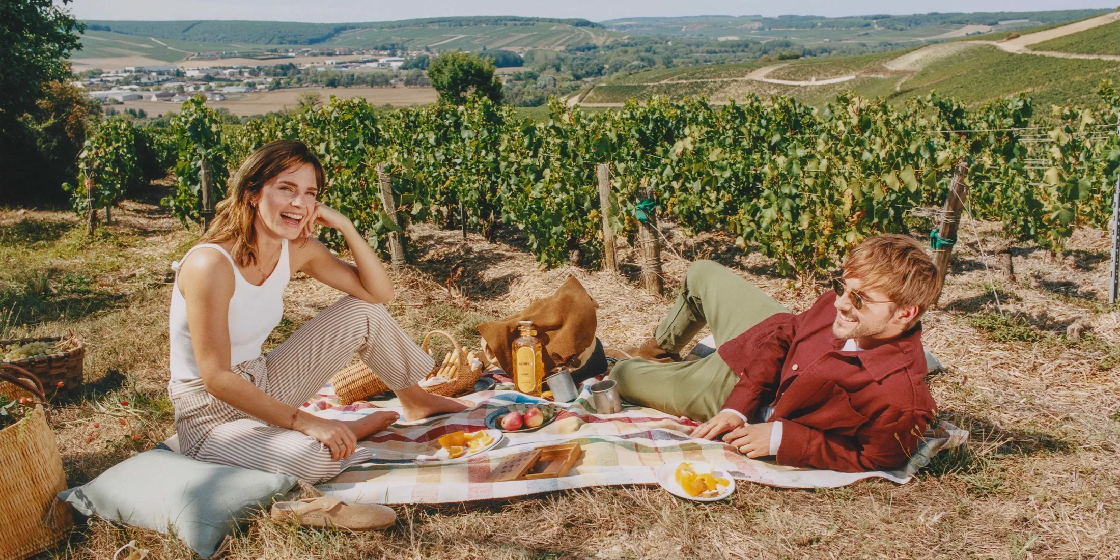 A smiling woman and man enjoy a picnic on a blanket in a vineyard, surrounded by food and drinks, with rolling green hills and farmland in the background under a clear sky.