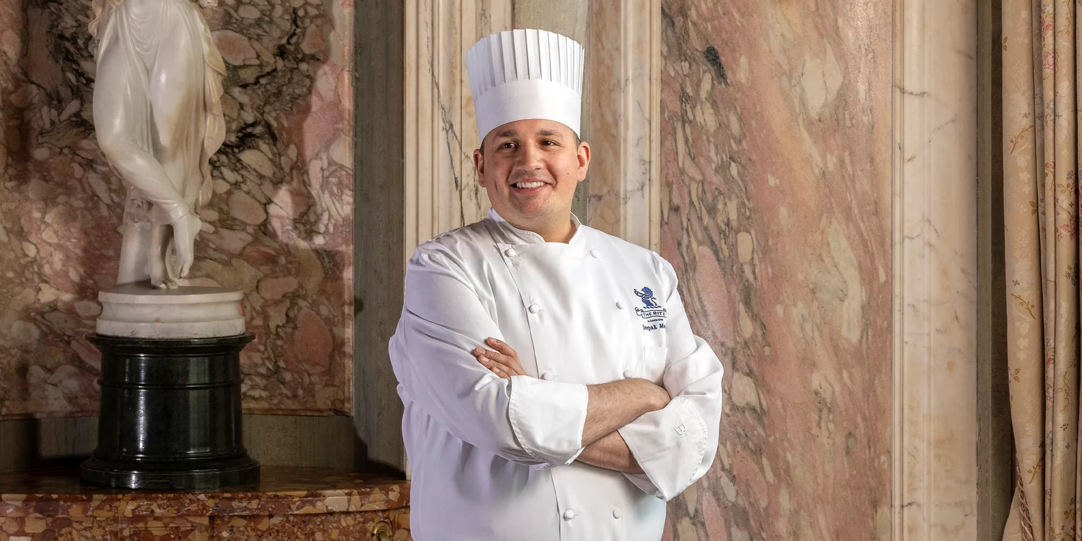 A smiling chef in a white uniform and tall chef hat stands with arms crossed in front of marble walls and a white statue in the background.