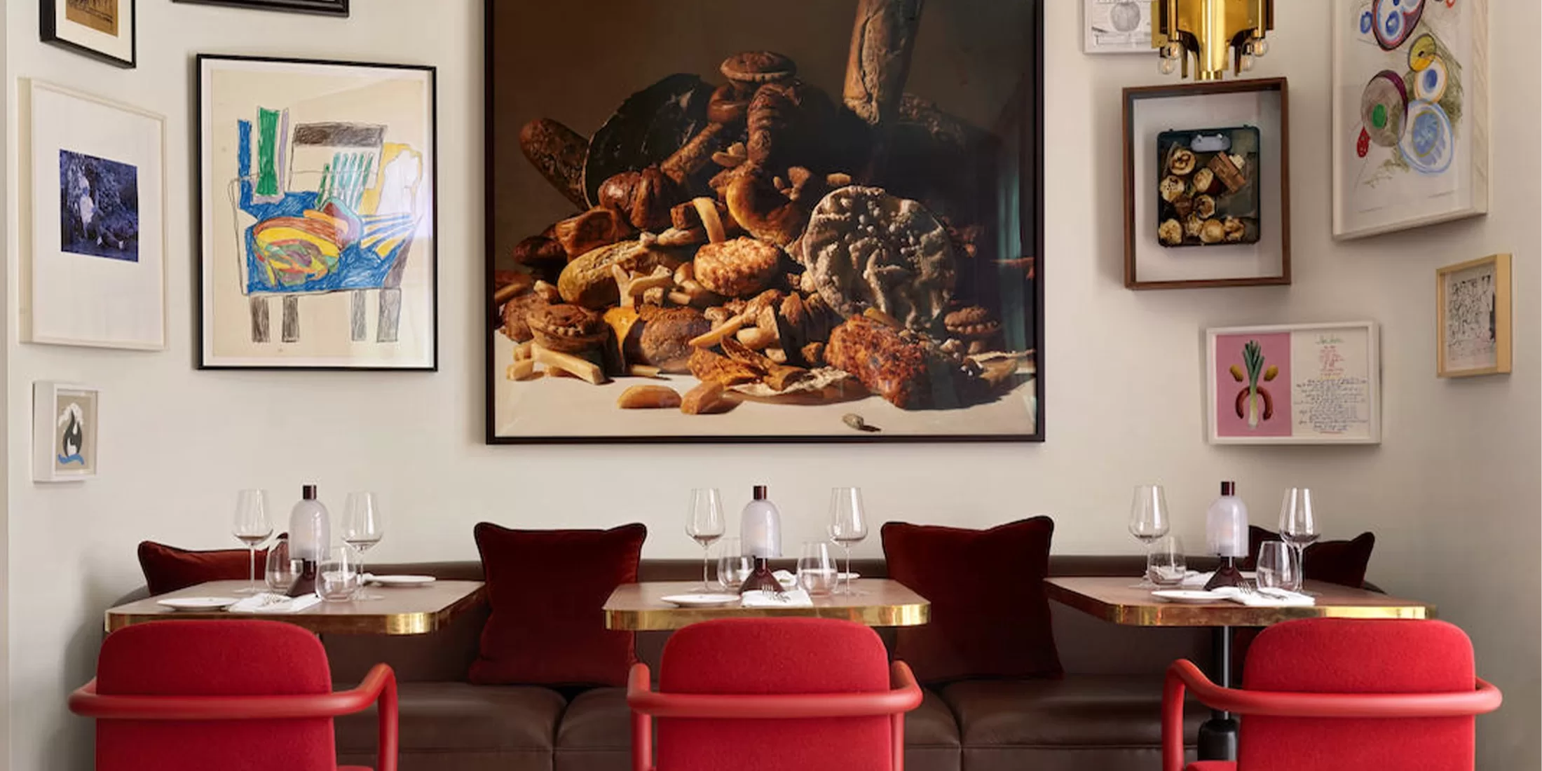 A cosy dining space with red chairs and a brown bench, set with wine glasses and napkins. The walls are adorned with eclectic framed artwork, including a large picture of assorted bread and mushrooms.