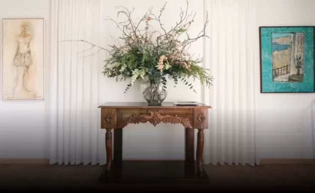 A wooden table features an ornate vase with a large floral arrangement of branches and greenery. It is set against white curtains, flanked by a sketch of a figure on the left and a framed blue artwork on the right.