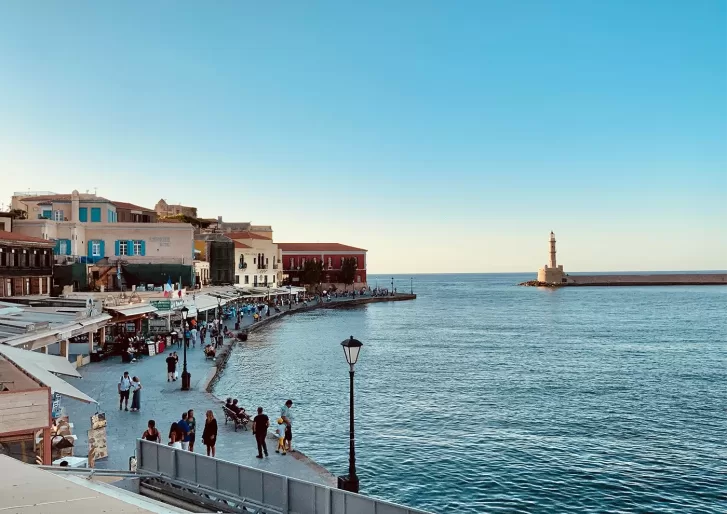 A waterfront promenade with people walking along cafes and shops, colorful buildings to the left, and a lighthouse on a distant pier under a clear blue sky beside calm sea waters.