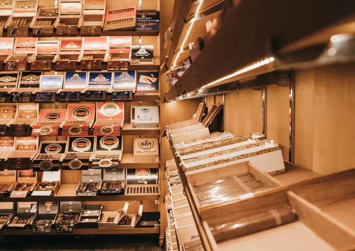 Wooden shelves display various boxes and rows of cigars in a well-lit humidor or cigar shop, with different brands and packaging visible.