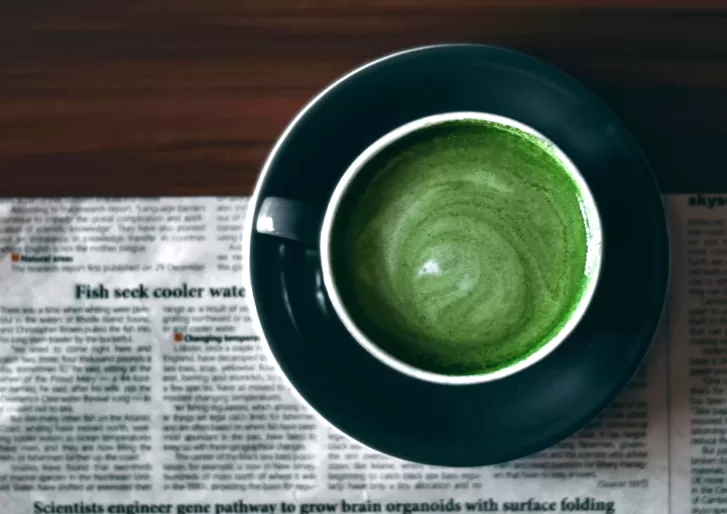A cup of green matcha latte seen from above, placed on a dark saucer and sitting on a newspaper on a wooden table. The latte forms a swirling pattern in the middle of the cup.