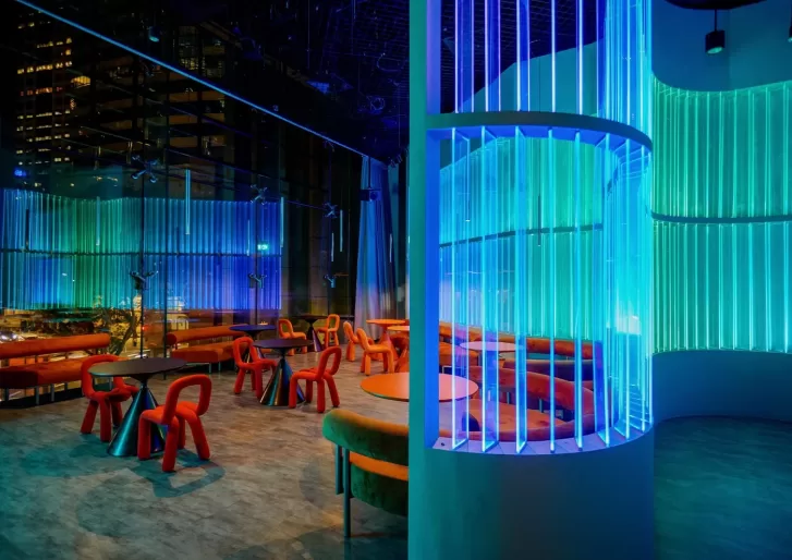 A modern, colorful lounge with curved benches, red chairs, and glowing blue and green vertical light bars, set against large windows overlooking a city at night.