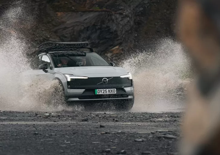 A white SUV with a roof rack drives through a large puddle, splashing water on a rocky, rugged road with mountains in the background. The cars headlights are on and the license plate reads OY25 NKO.