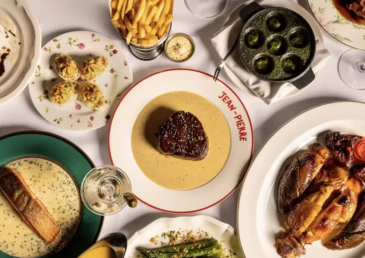 A table set with steak in creamy sauce, roast chicken, salmon, asparagus, escargot, fries, deviled eggs, a glass of white wine, and a small dish of butter, on elegant plates, one labeled “Jean Pierre.”.