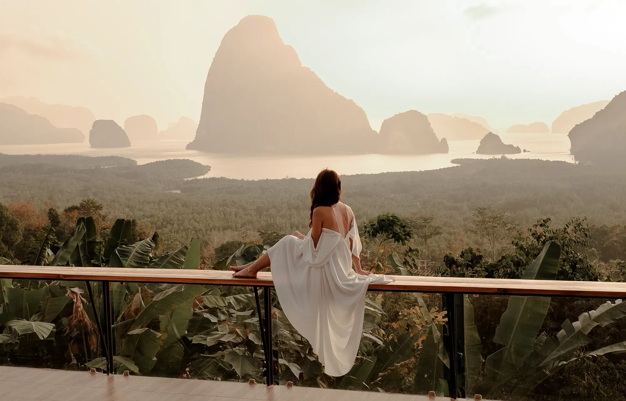 A woman in a white dress sits on a wooden railing, overlooking lush greenery and dramatic limestone cliffs rising from the water at sunrise or sunset.