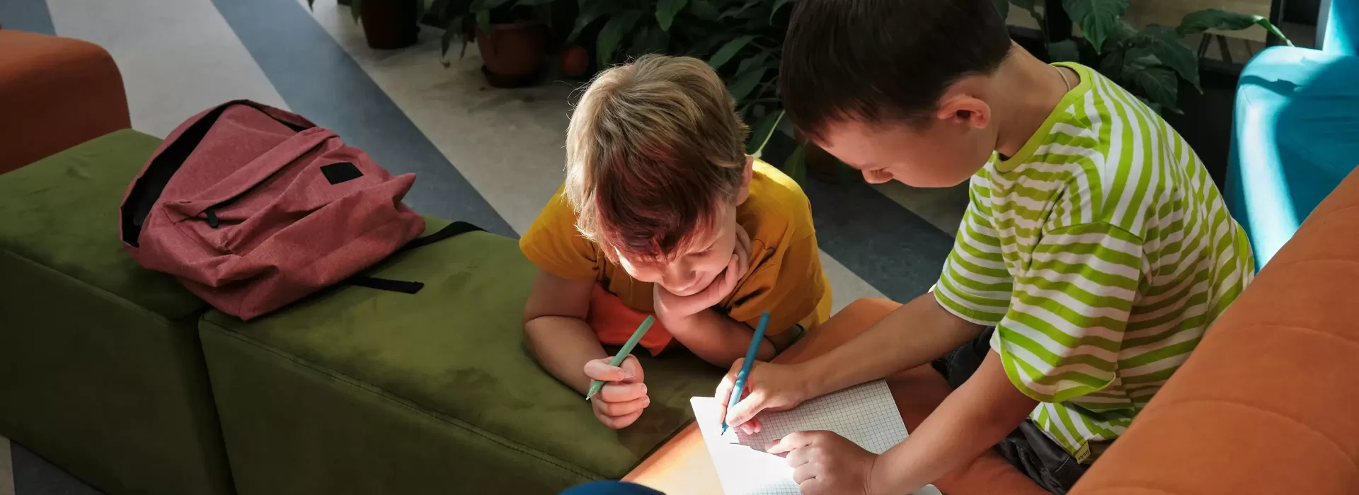 Two young boys writing on a notebook together, illuminated by sunlight. One wears a yellow shirt and the other a green striped shirt. A pink backpack rests on a green cushion beside them. Plants are in the background.
