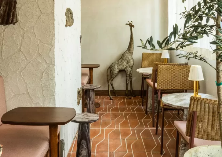 A cozy cafe corner with tiled floors, wooden tables, wicker chairs, potted plants, and a decorative giraffe statue against a cream-colored wall.