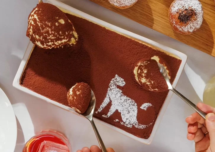 A rectangular tiramisu dusted with cocoa powder features a powdered sugar silhouette of a cat. Two hands with spoons are scooping servings from the dessert. Other pastries and a pink drink are partially visible nearby.
