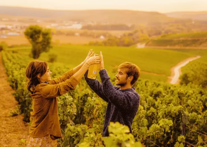 Two people stand among lush green vineyard rows, both smiling and reaching up to hold a wine bottle together, with golden sunlight and rolling hills in the background.