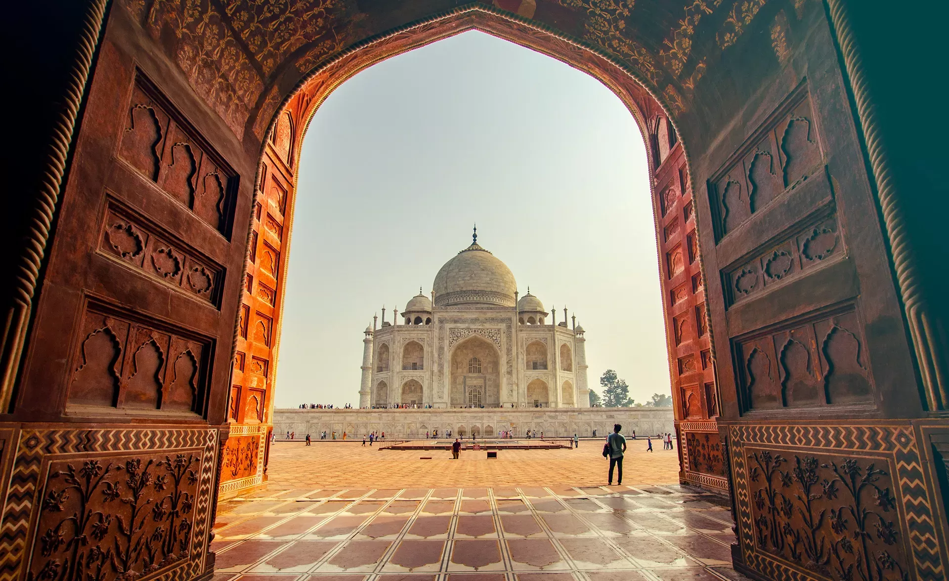 View of the Taj Mahal through a grand archway. The sunlit white marble mausoleum stands tall against a clear sky, with people walking on the red stone courtyard in the foreground. Intricate patterns adorn the archway.